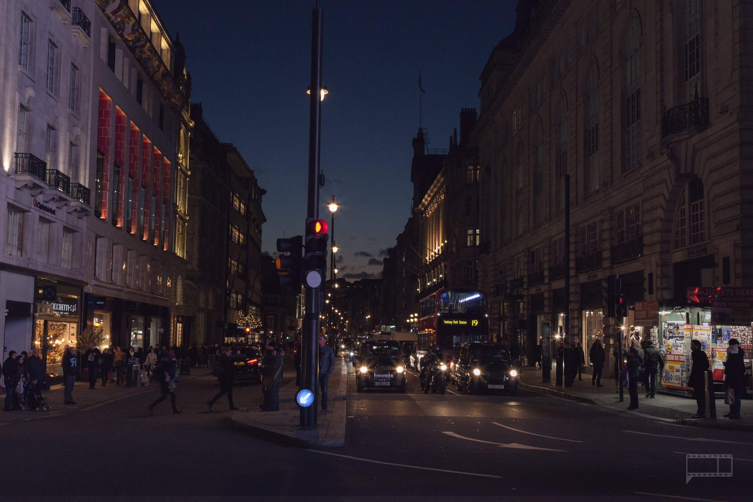 City street at dusk with cars, buses, pedestrians, and illuminated buildings.