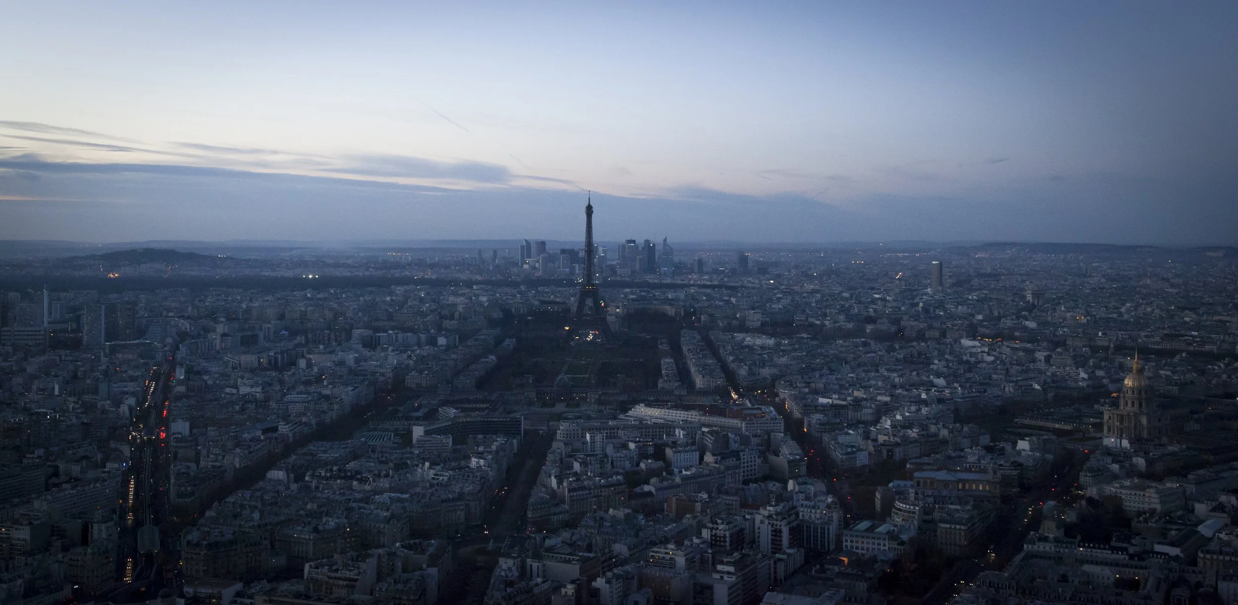 An aerial view of Paris at dusk, featuring the Eiffel Tower illuminated and city streets with traffic lights visible.