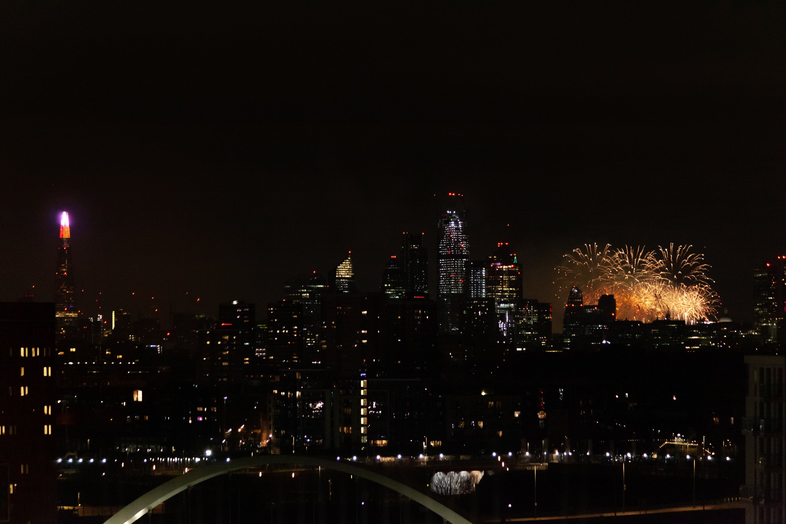 Nighttime cityscape with fireworks in the background, tall illuminated skyscrapers, and a lit-up bridge or structure in the foreground.