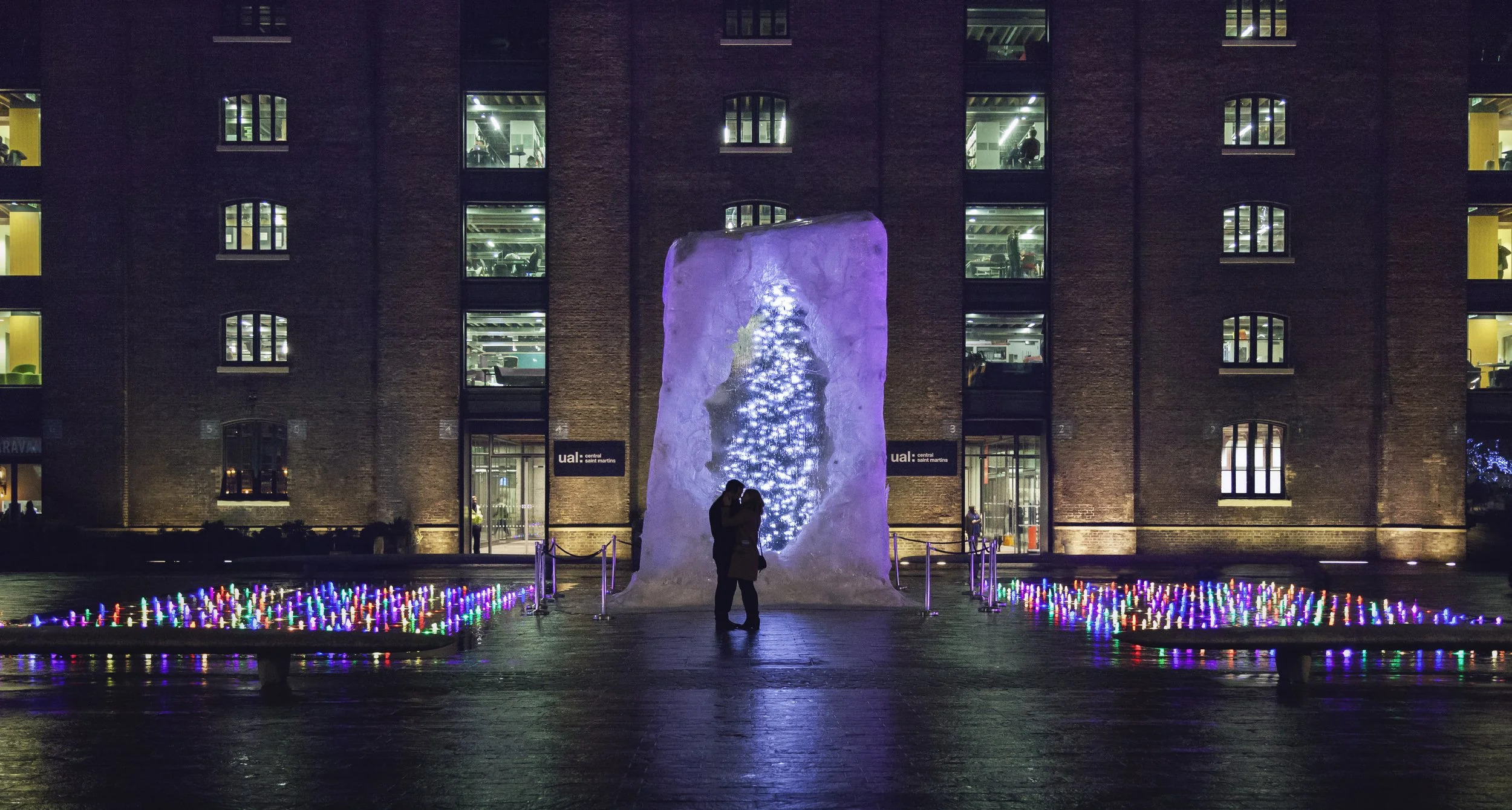 Night scene of a large ice sculpture with a Christmas tree inside, illuminated with lights, standing in front of a dark brick building with lit windows, and decorated with colorful lights on the ground.