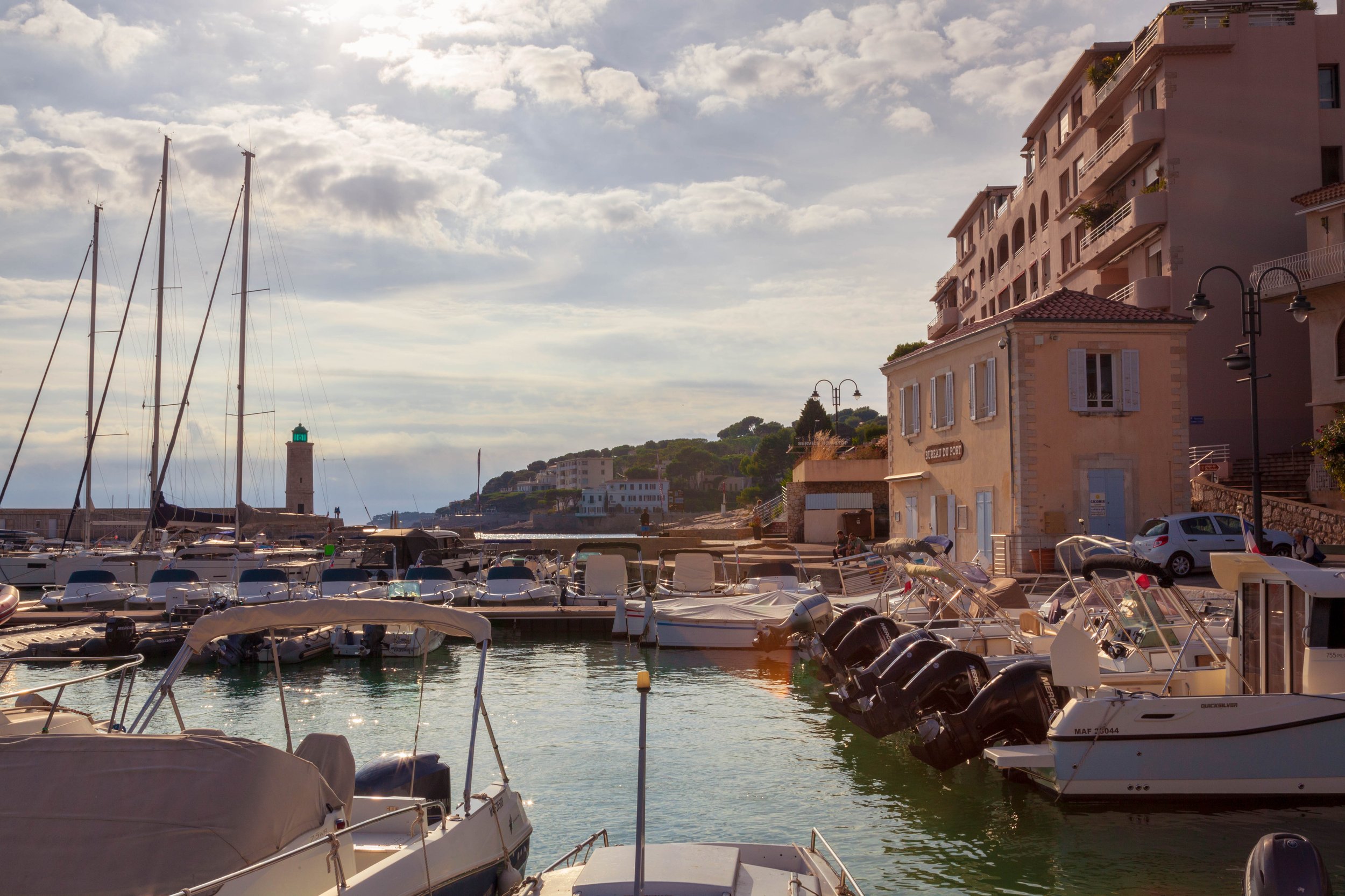 A serene marina filled with boats and yachts, with a lighthouse in the background, beside pastel-colored buildings under a partly cloudy sky.