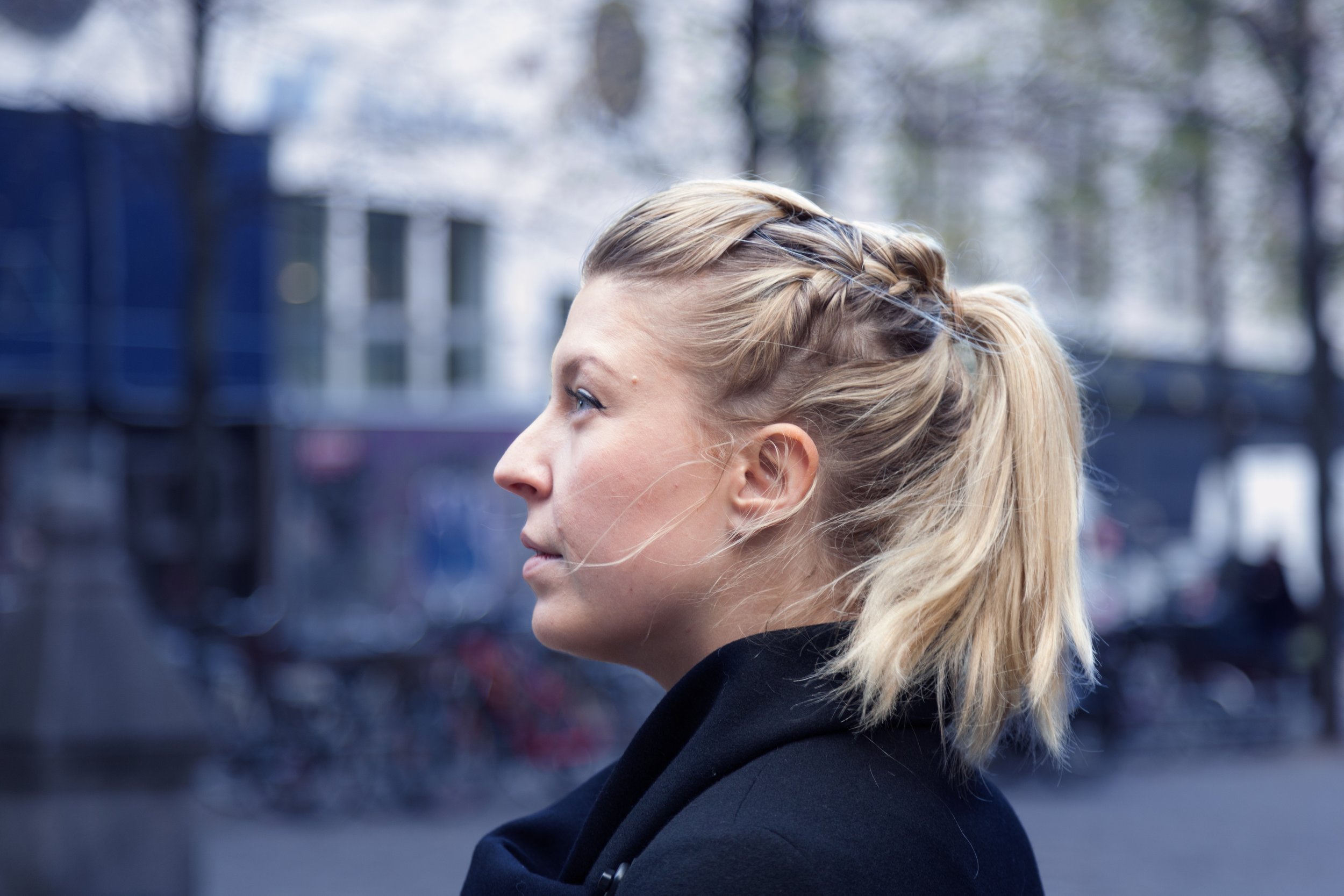 Profile of a blonde woman with a braid in her hair, looking to the left, outside in an urban area with blurred buildings and trees in the background.