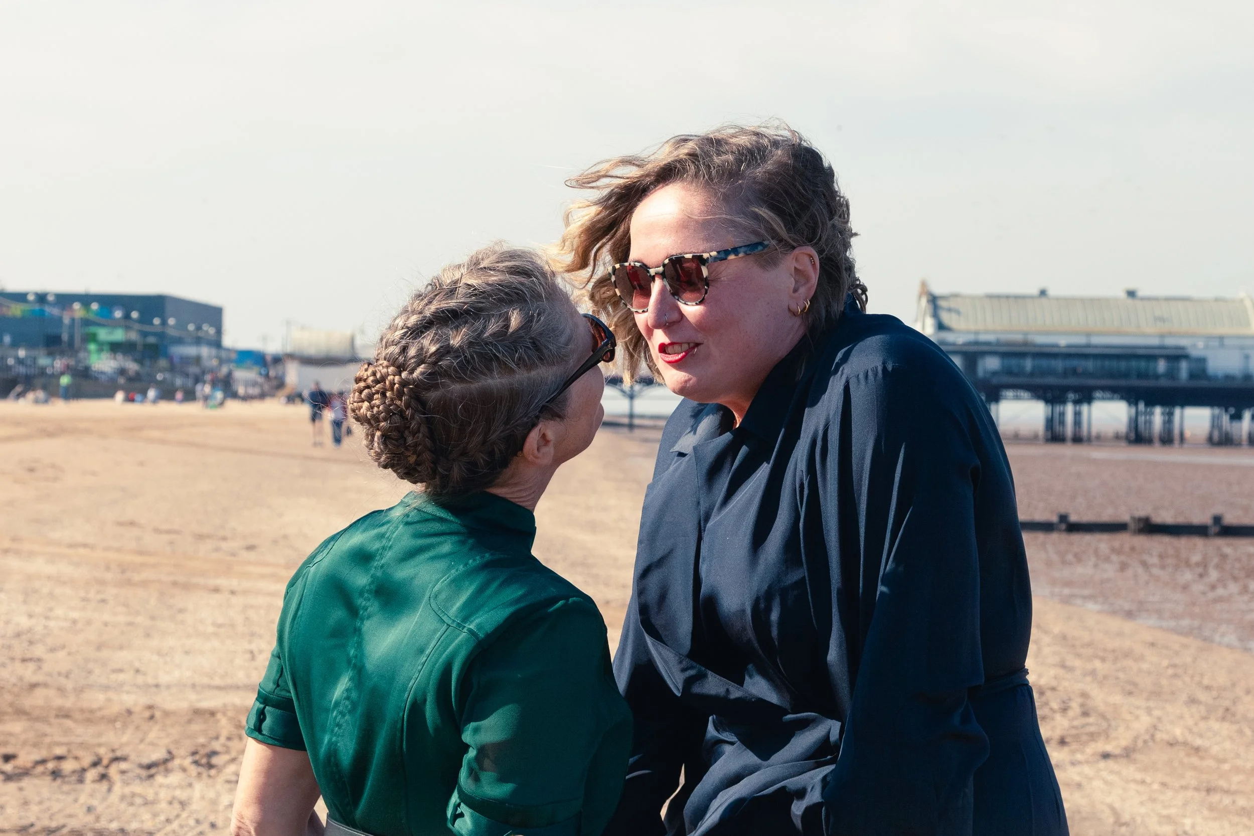 Two women facing each other and talking on a sandy beach with a pier in the background, one wearing a green dress and the other a nay dresst with sunglasses.