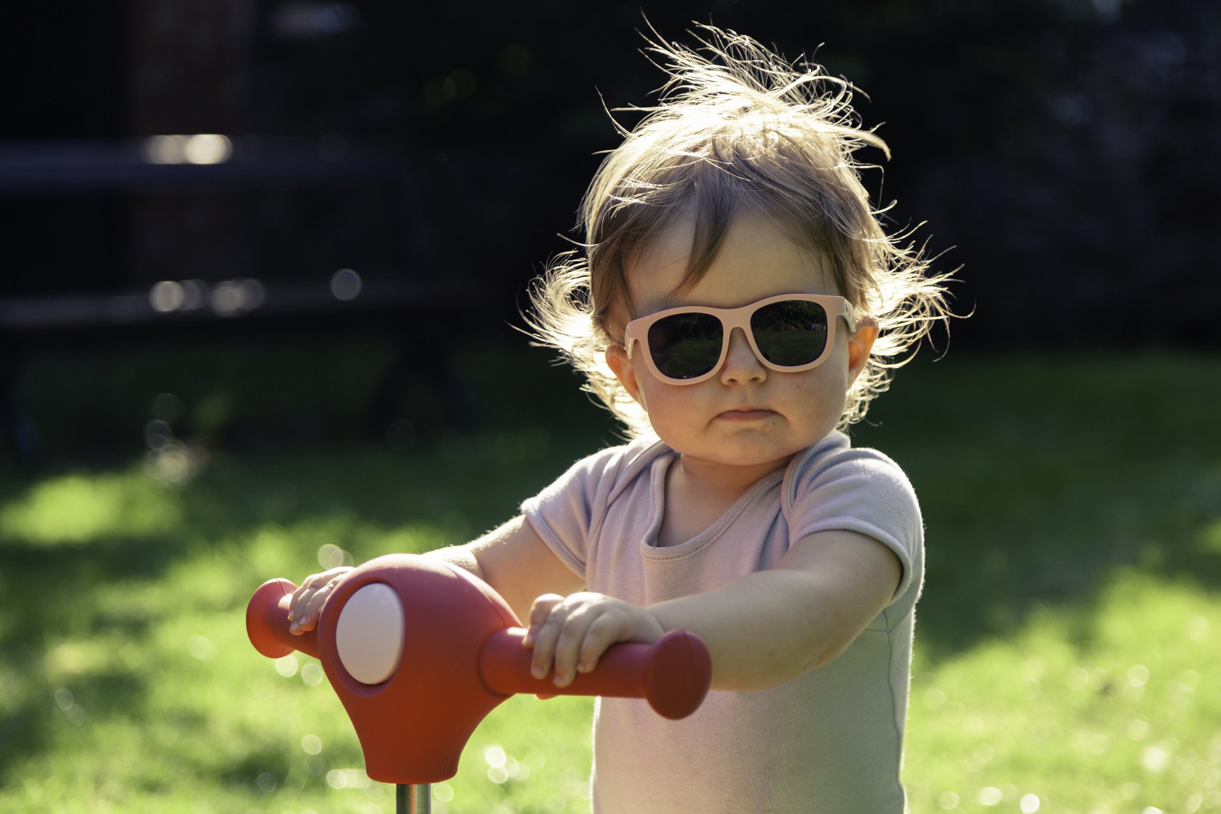 A young child wearing pink sunglasses and a light-colored shirt, standing outdoors on a sunny day, holding onto a red and white scooter.