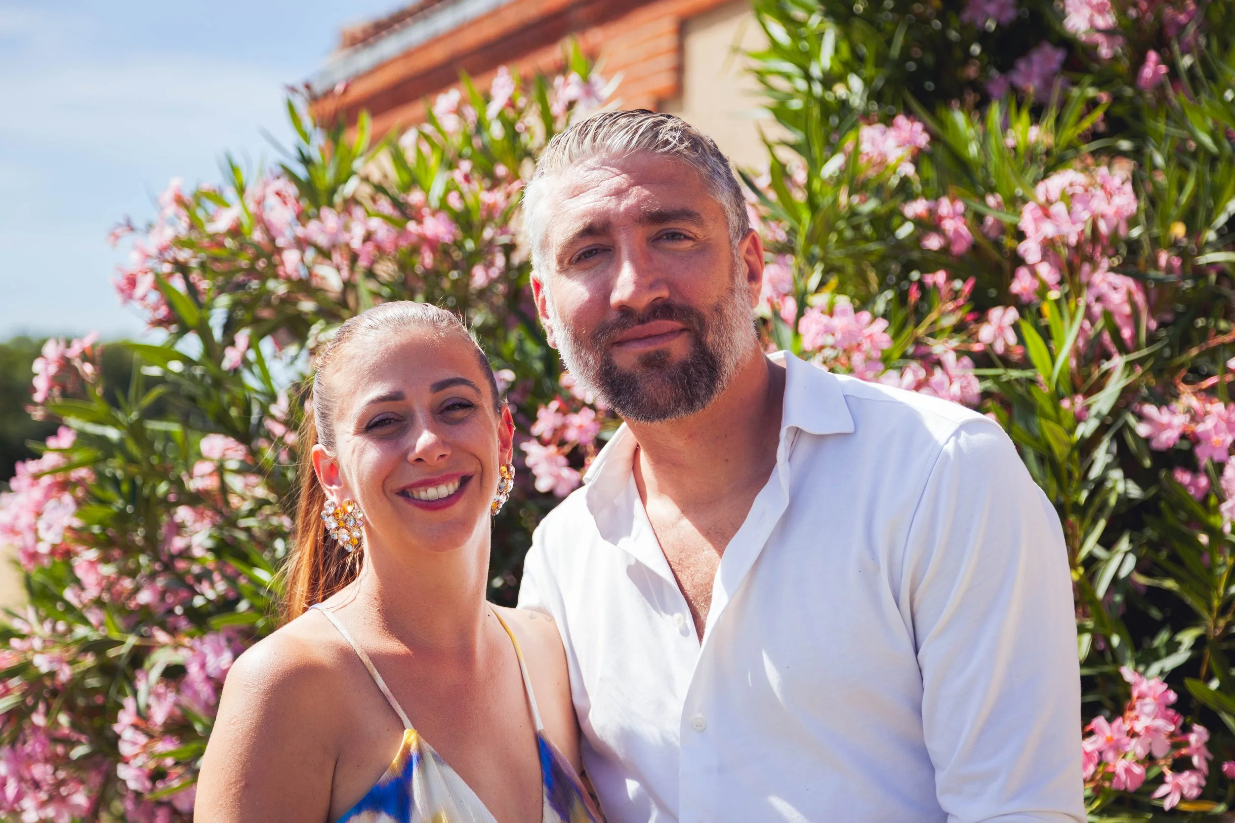 A smiling couple standing outdoors near pink flowering bushes and a house with a tiled roof on a sunny day.