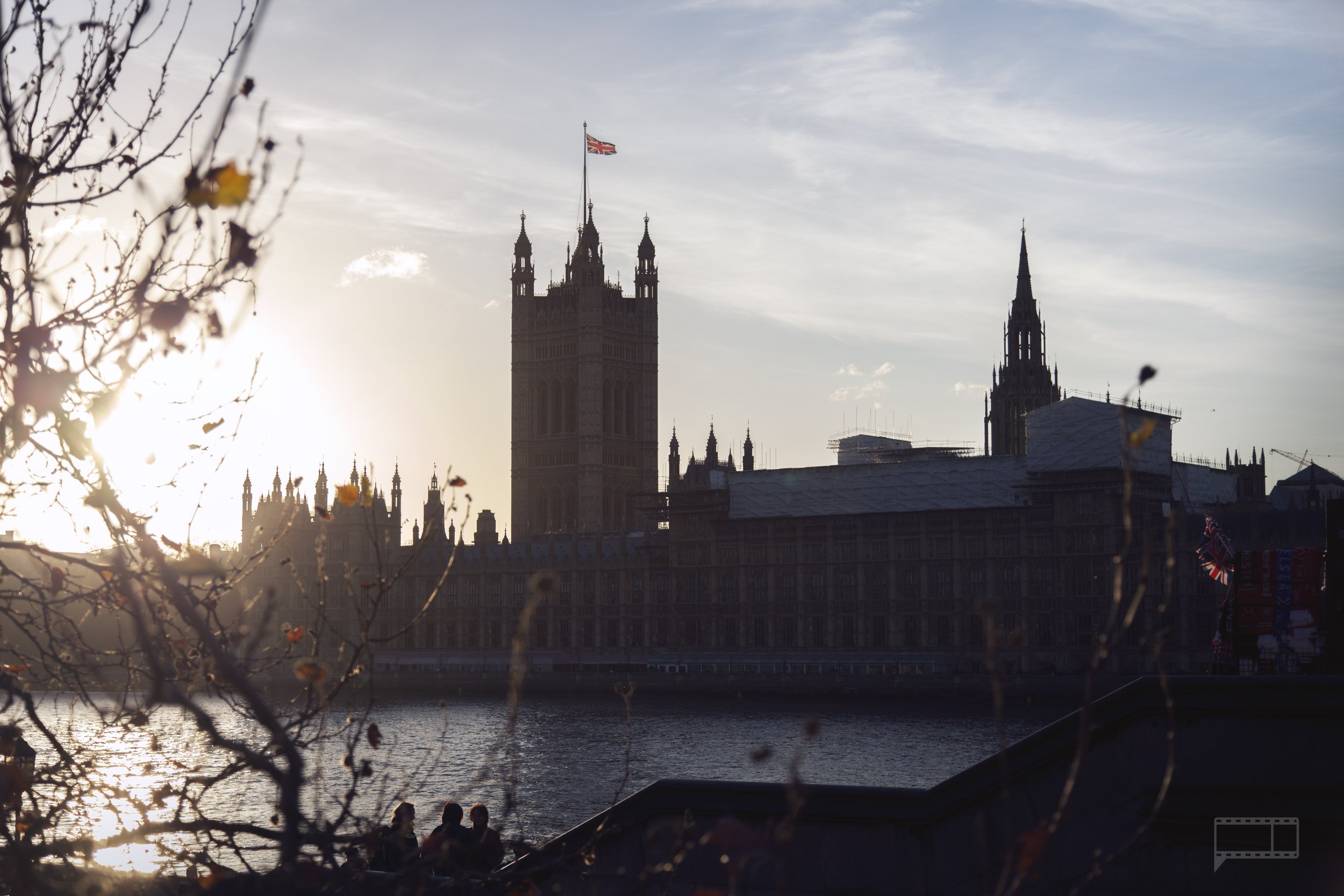 View of the Houses of Parliament and Big Ben in London with the river and trees in the foreground during sunset.
