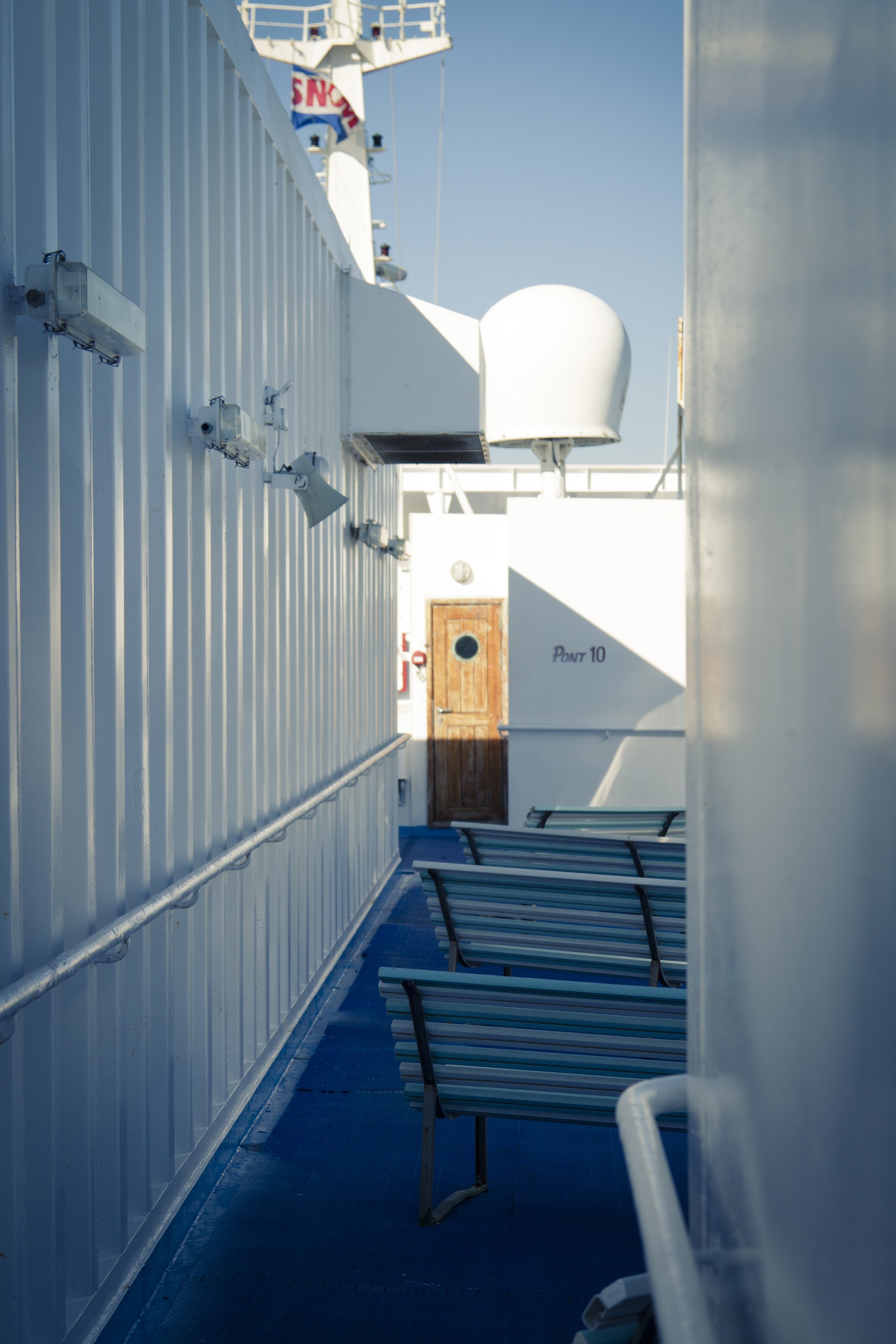 View along the deck of a cruise ship with white walls, benches, and various electronic and communication equipment, leading to a wooden door at the end of the corridor.