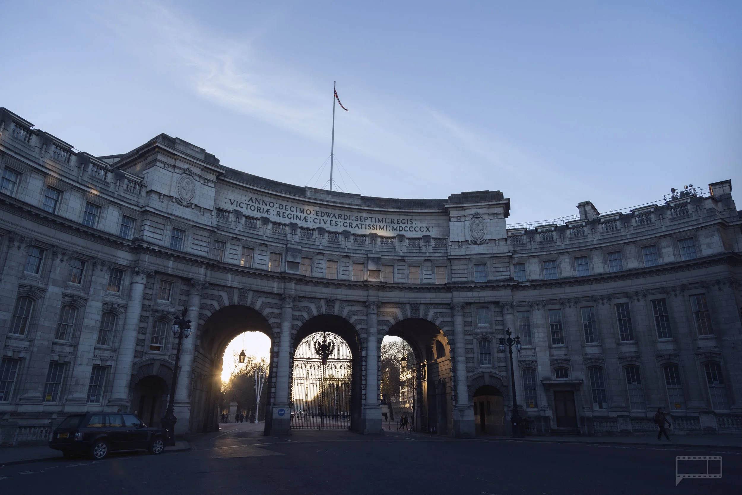 View of a grand, curved building with three arches, an ornate gate, and a flagpole with a flag on top. The sun is setting behind the building.