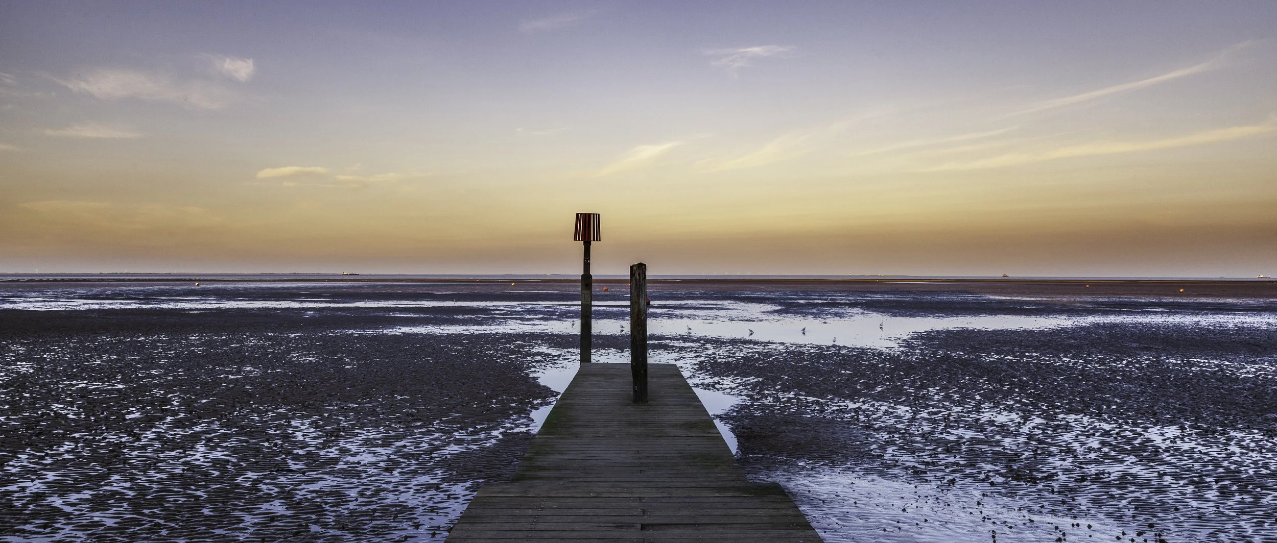 A wooden pier extending into a tidal flat during sunset, with a red navigation marker and distant ships on the horizon.