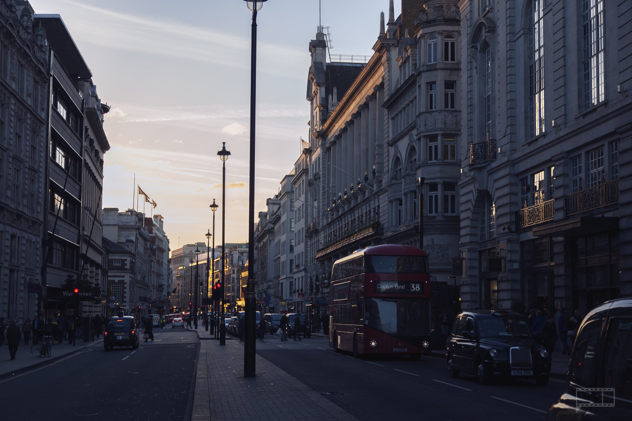 City street scene at sunset with historic buildings, a red double-decker bus, cars, pedestrians, and streetlights.