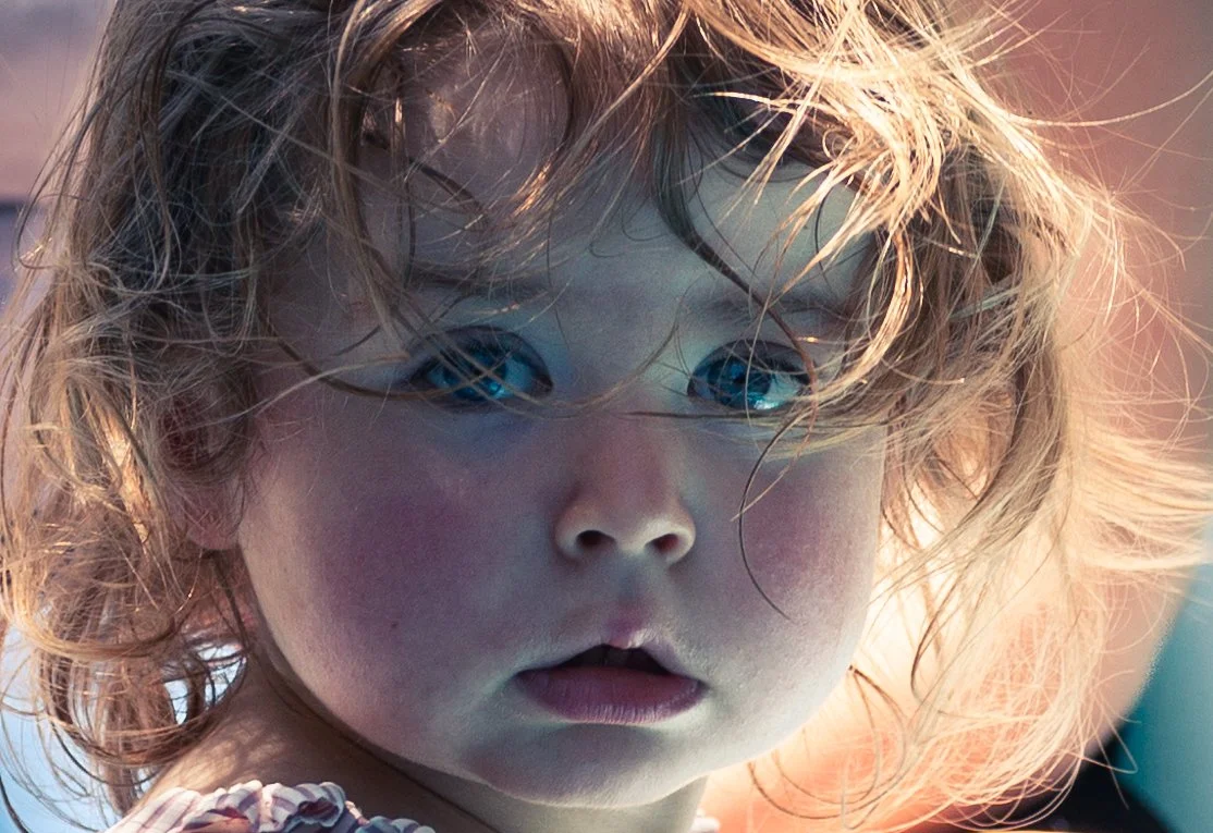 Close-up of a young girl with curly blonde hair and blue eyes, looking at the camera with a surprised expression.