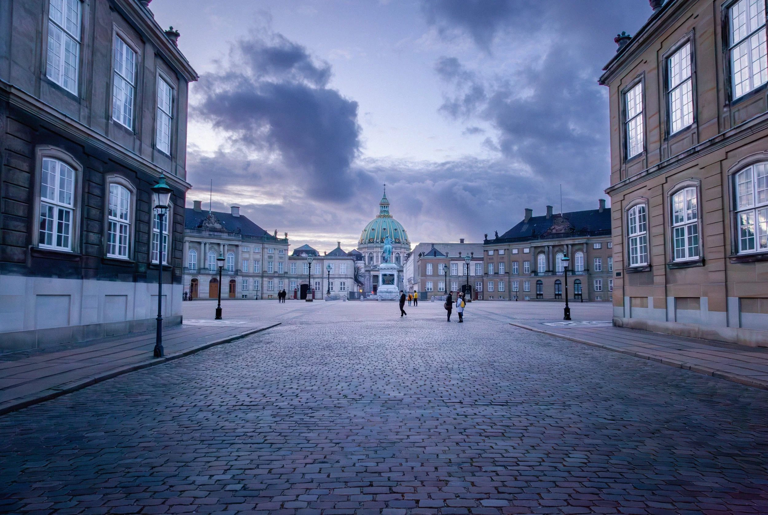 A cobblestone square surrounded by historic buildings with large windows, lamp posts, and a central statue with a dome and statue on top in the background, under a cloudy sky at dusk.