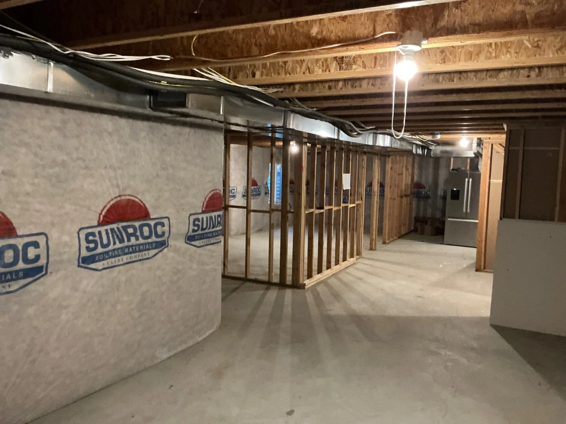 Interior view of a basement under construction, showing exposed wooden framing, insulation with 'Sunroc Building Materials' logo, and bare light bulbs hanging from the ceiling.