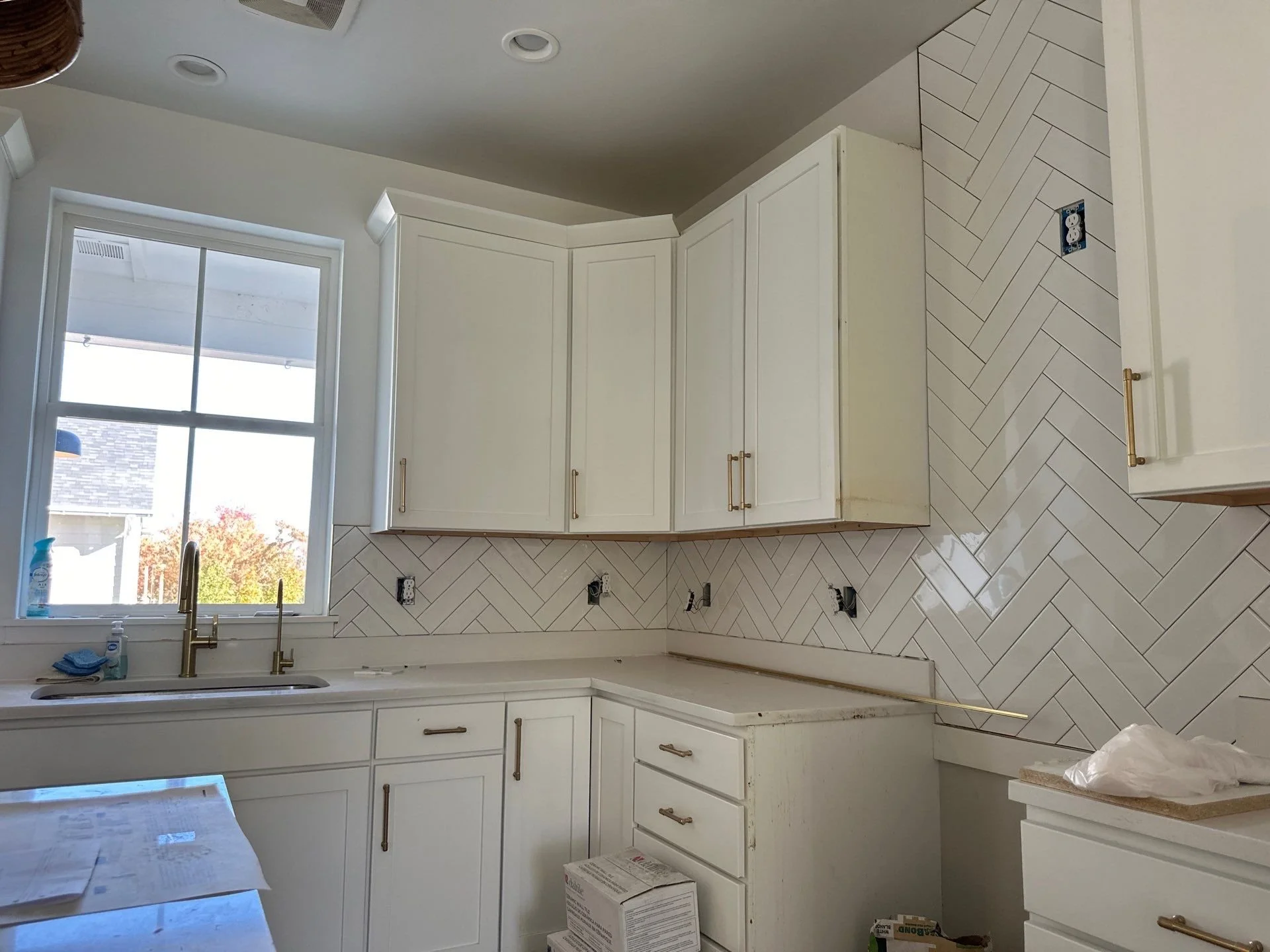 Kitchen with white cabinets, a large window above the sink, and a herringbone tile backsplash. Some electrical outlets are missing faceplates, and there are construction materials on counters and the floor.
