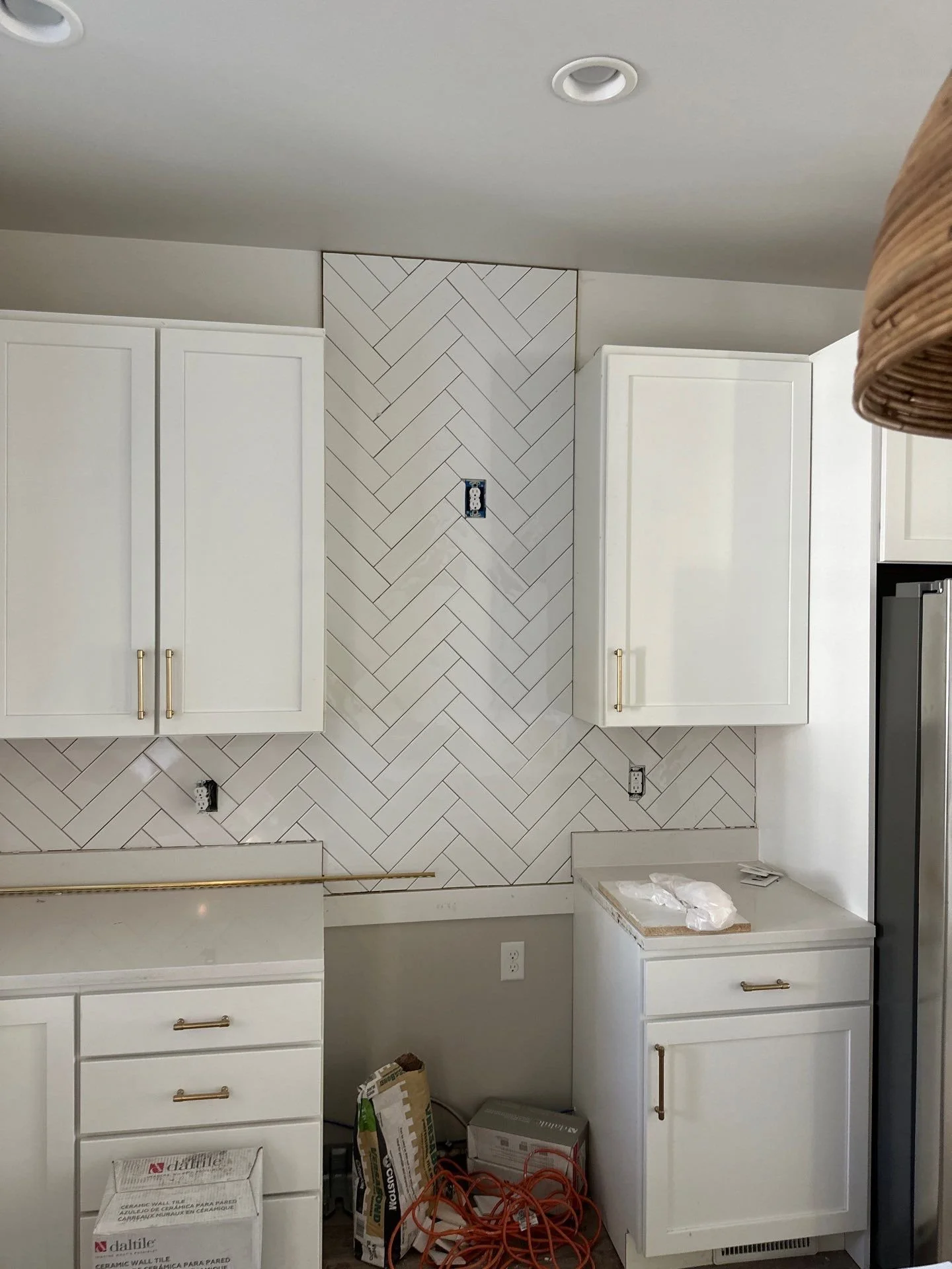Kitchen with white cabinets and a white herringbone tile backsplash. Cabinets lack hardware, and there are electrical outlets and boxes without covers. Construction materials and tools are on the lower cabinets and floor.