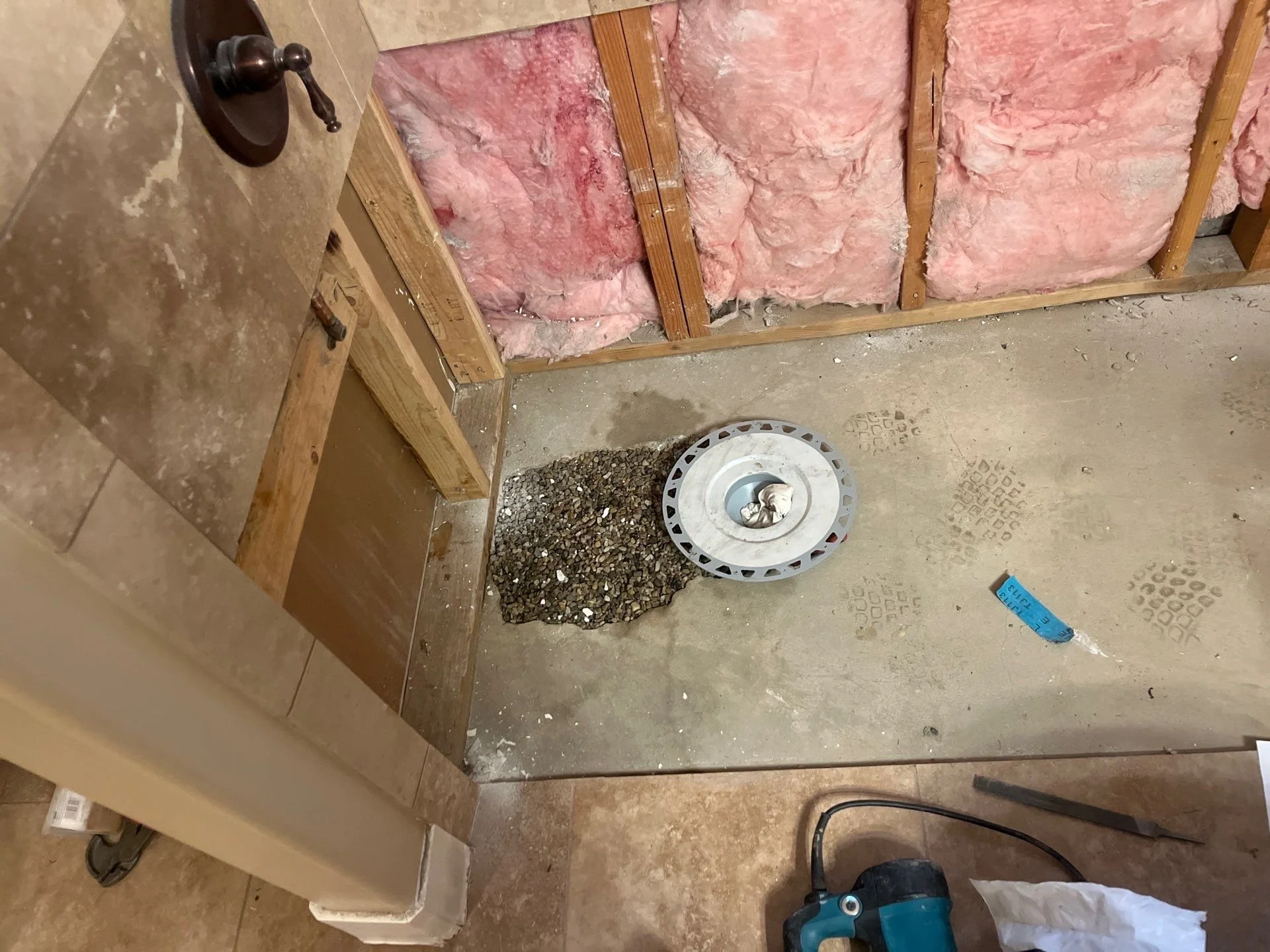 Bathroom renovation in progress with exposed pink insulation, a pile of gravel, a floor drain, and construction tools.