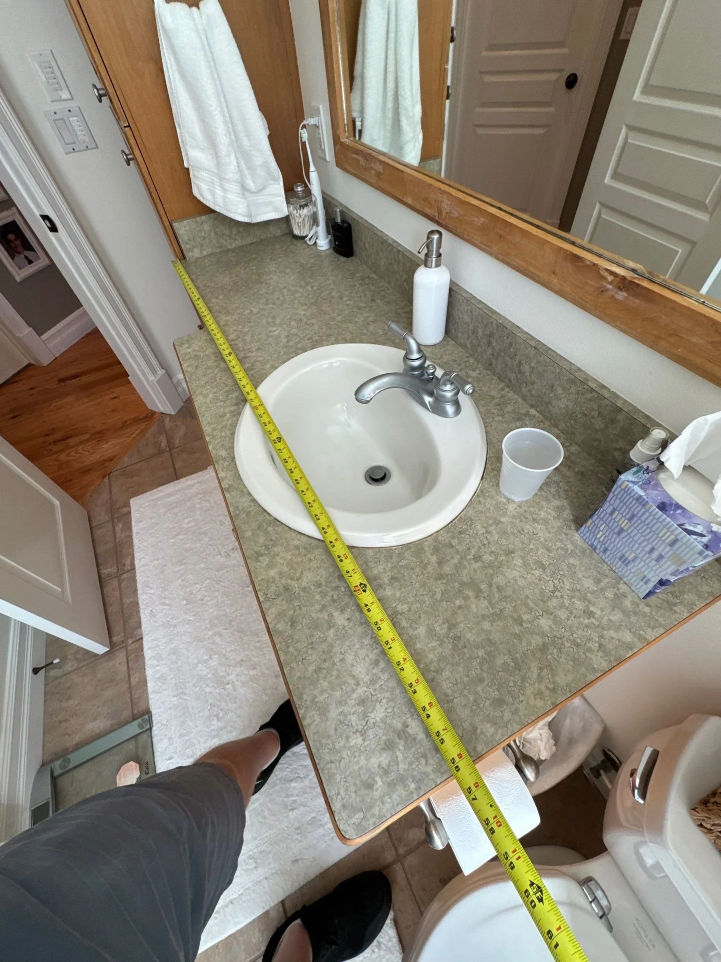 Bathroom countertop with a round sink, soap dispenser, tissue box, and cup, with a mirror and towels visible in the reflection, measuring tape stretched across the counter.