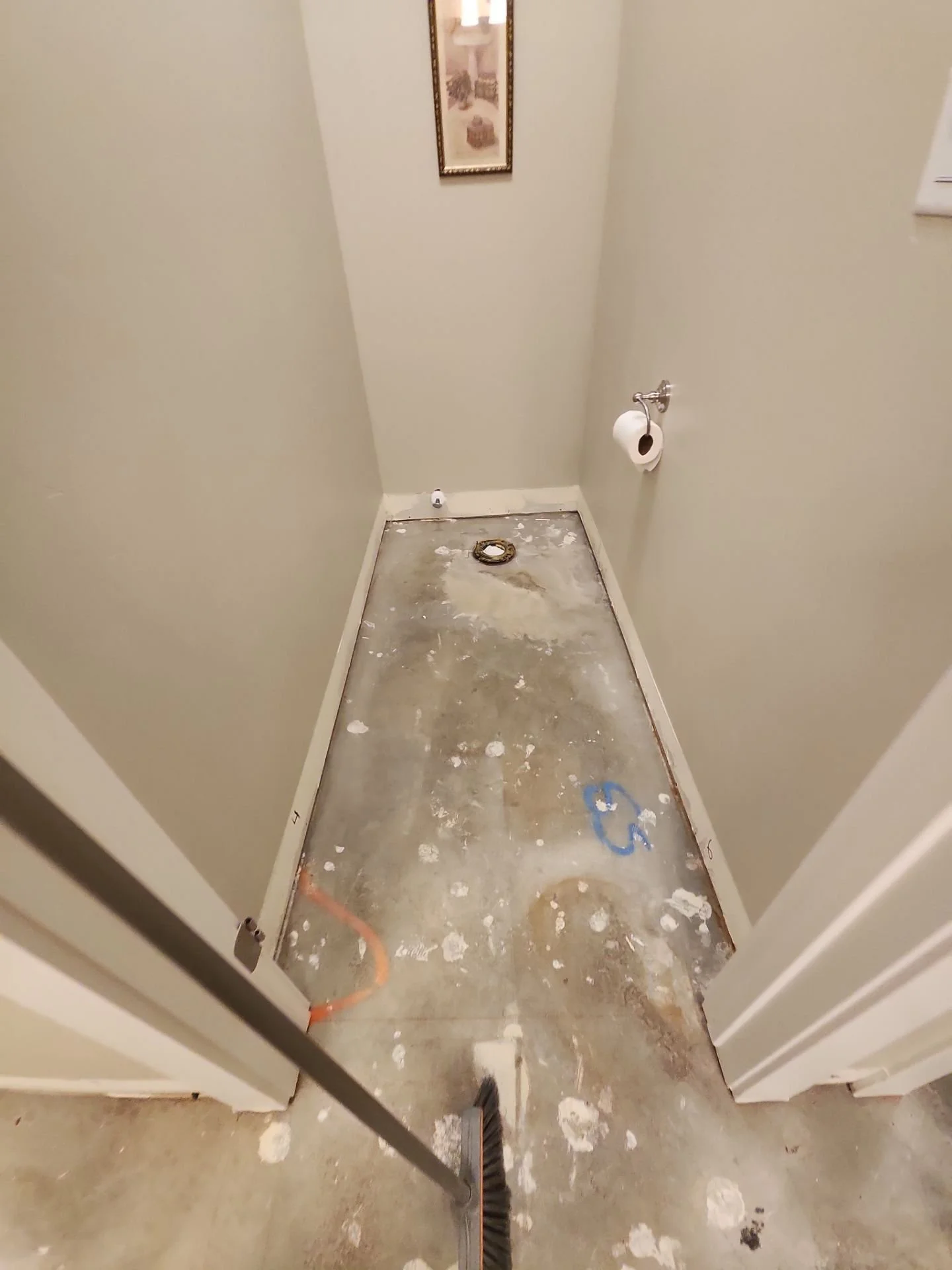 A small bathroom with the floor removed for renovation, showing exposed subfloor and plumbing. There is a toilet paper roll on a wall-mounted holder, a framed picture on the wall, and construction tools on the floor.