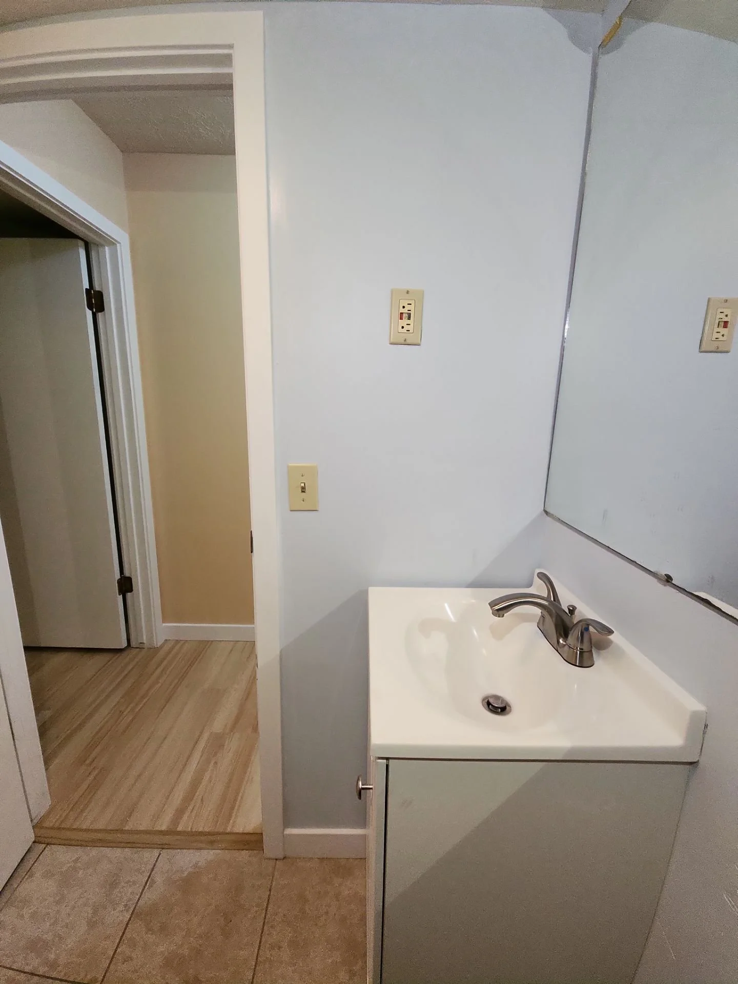 Small bathroom sink with faucet, mirror, electrical outlets, and open door leading to another room with wood flooring.