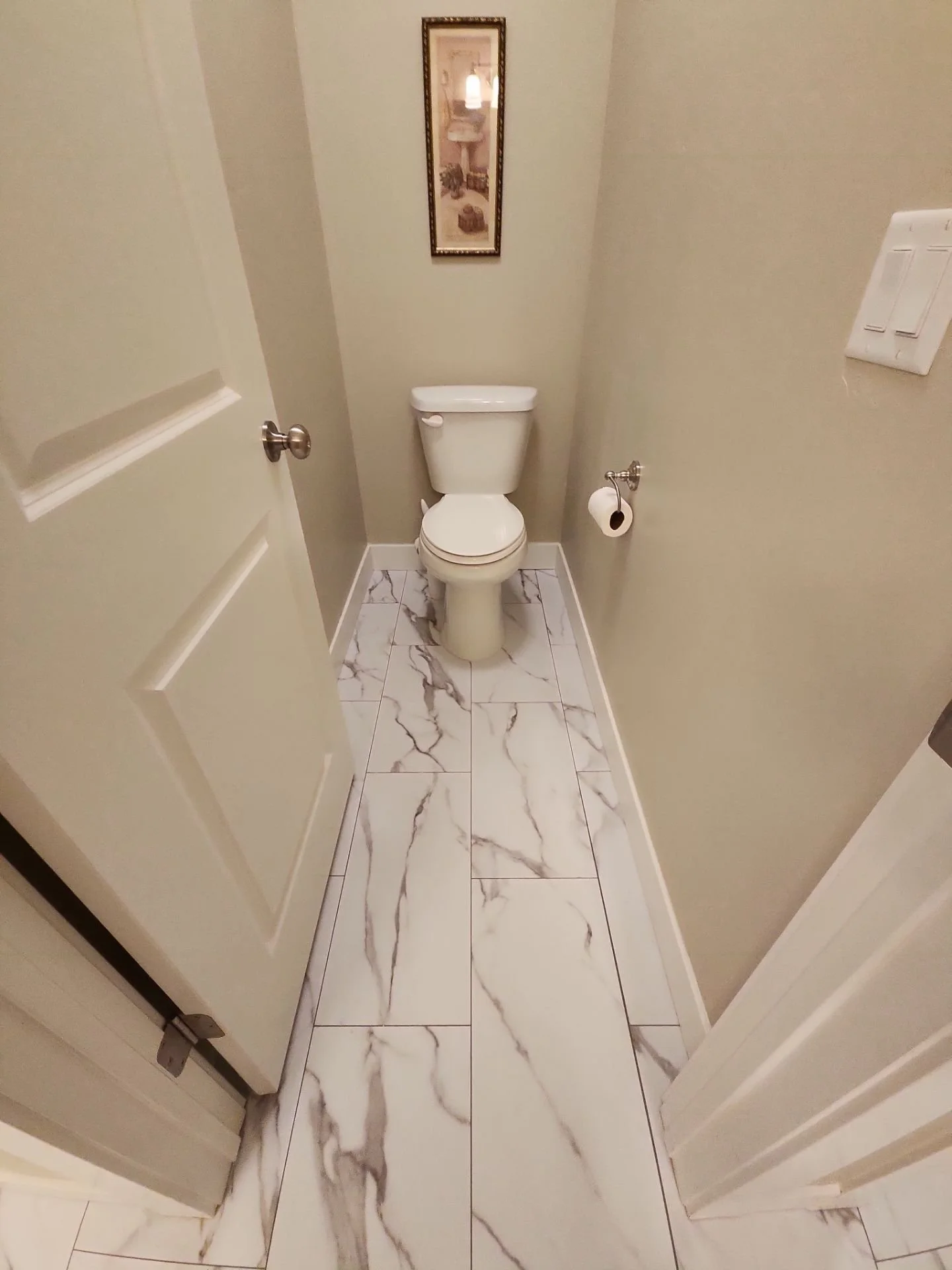 A small bathroom with marble floor tiles, a toilet, a wall mirror, a toilet paper roll, and a light switch.
