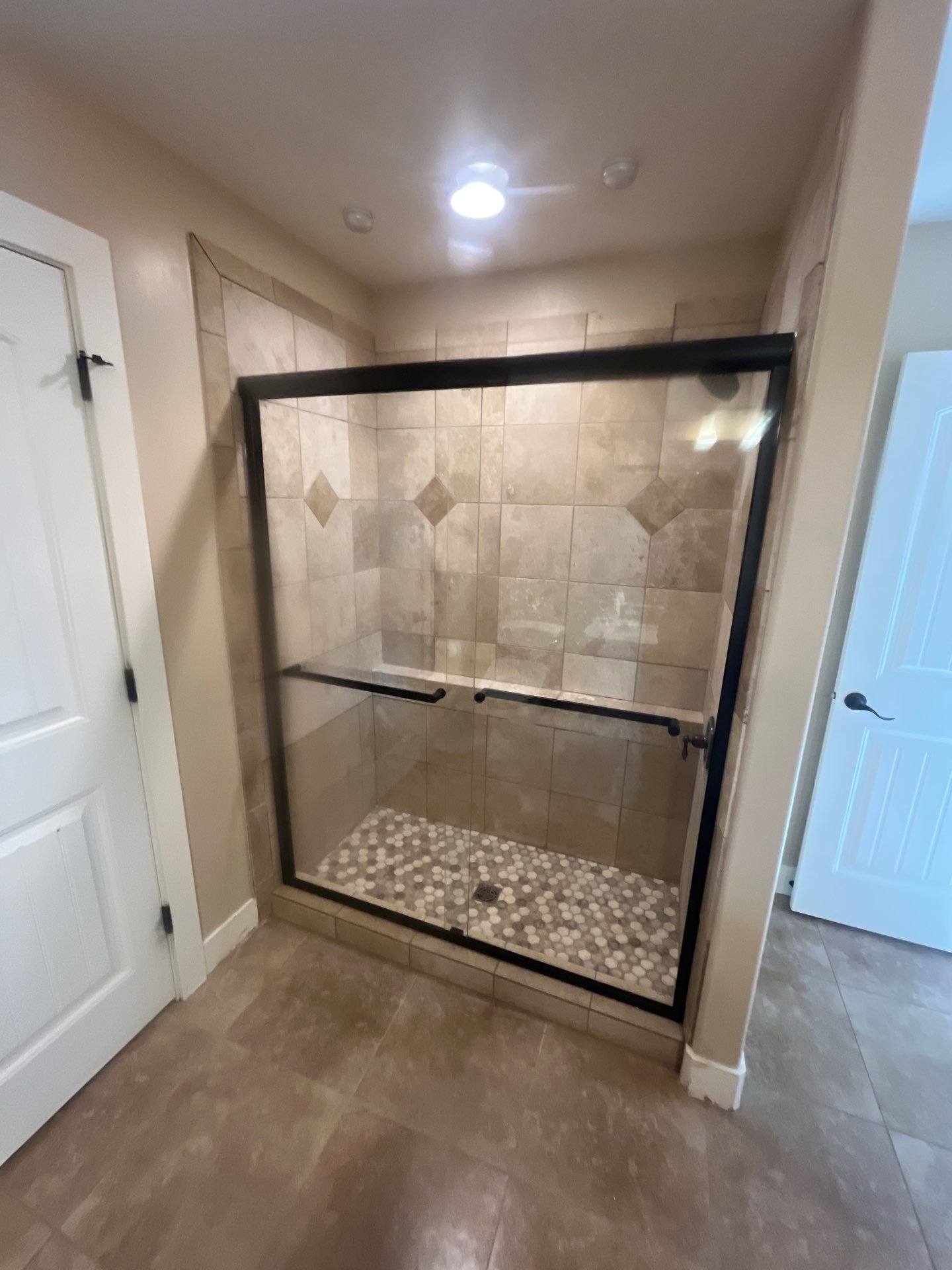 Bathroom shower with glass enclosure, beige tiled walls with diamond accents, pebble tile floor, and corner grab bars.