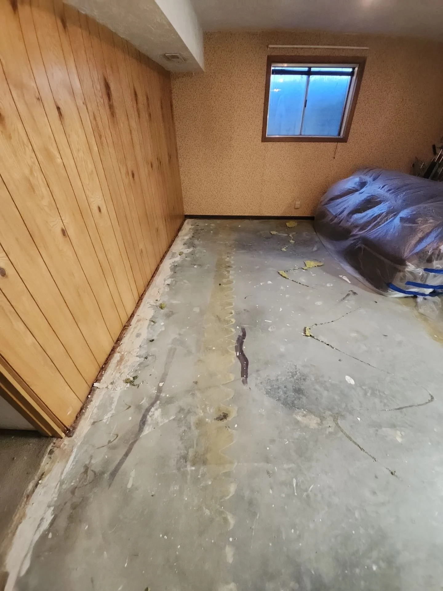 A room with wooden paneling on one wall, a window with a blue hue outside, and a partially visible bed covered with plastic, with the floor removed showing the concrete subfloor.