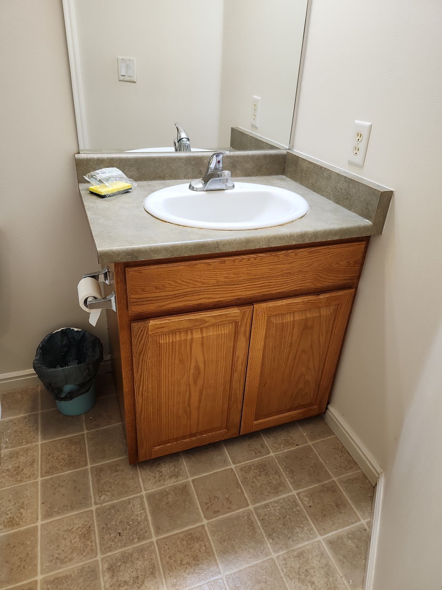 Bathroom vanity with a round white sink, a faucet, a mirror, a yellow sponge, and a trash can with a plastic liner.