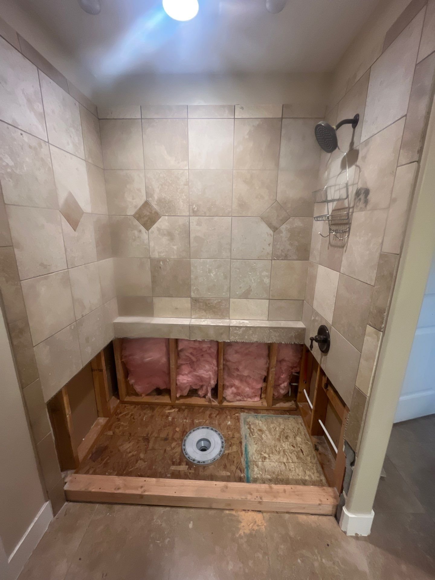 A bathroom shower with beige tiles, a built-in bench, and a showerhead. The lower portion is unfinished with exposed plumbing, insulation, and wood framing.