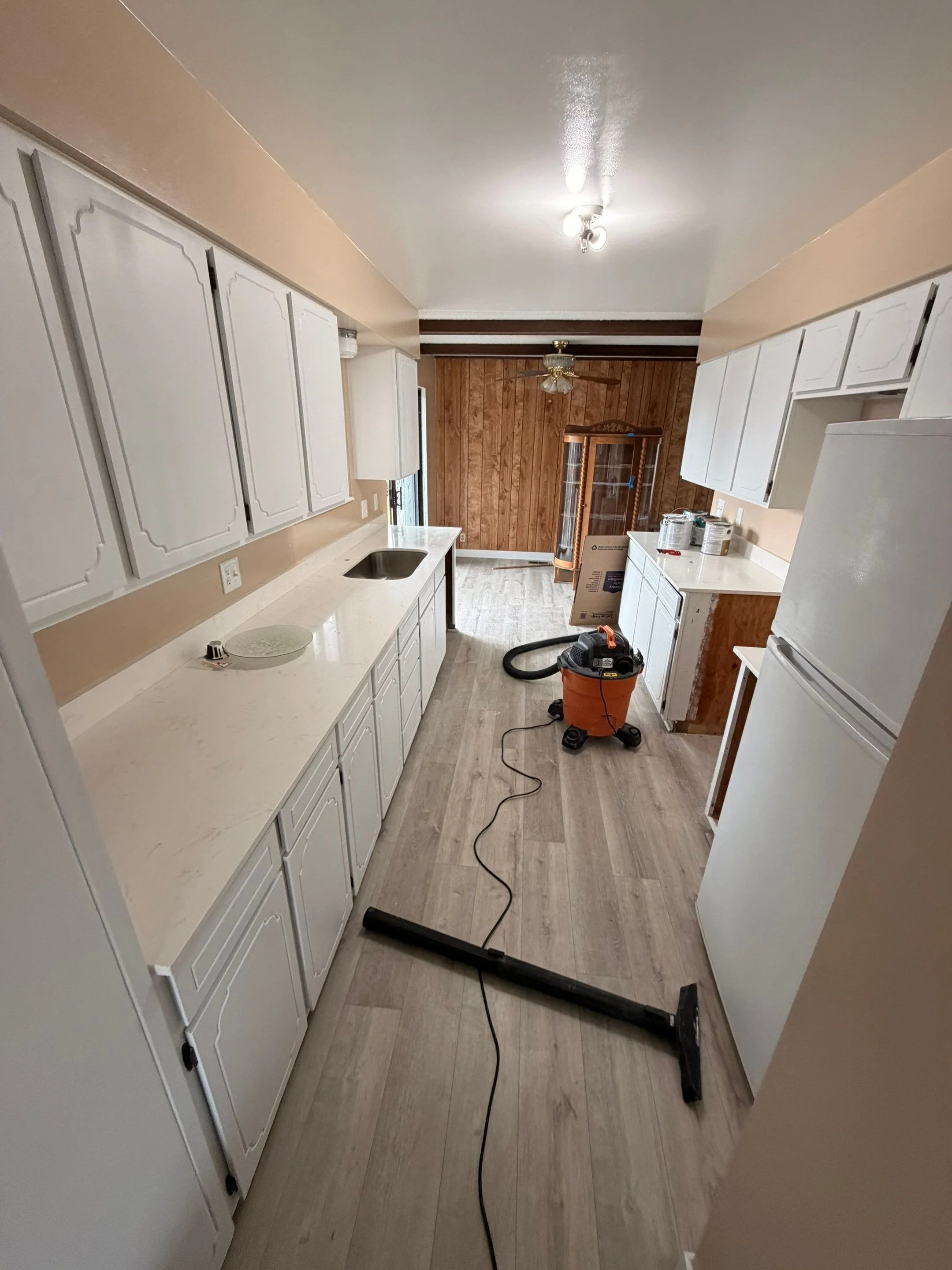 Kitchen with white cabinets, beige walls, and a wooden wall at the far end. Cleaning tools and equipment, including a vacuum with a hose and a box, are on the floor.