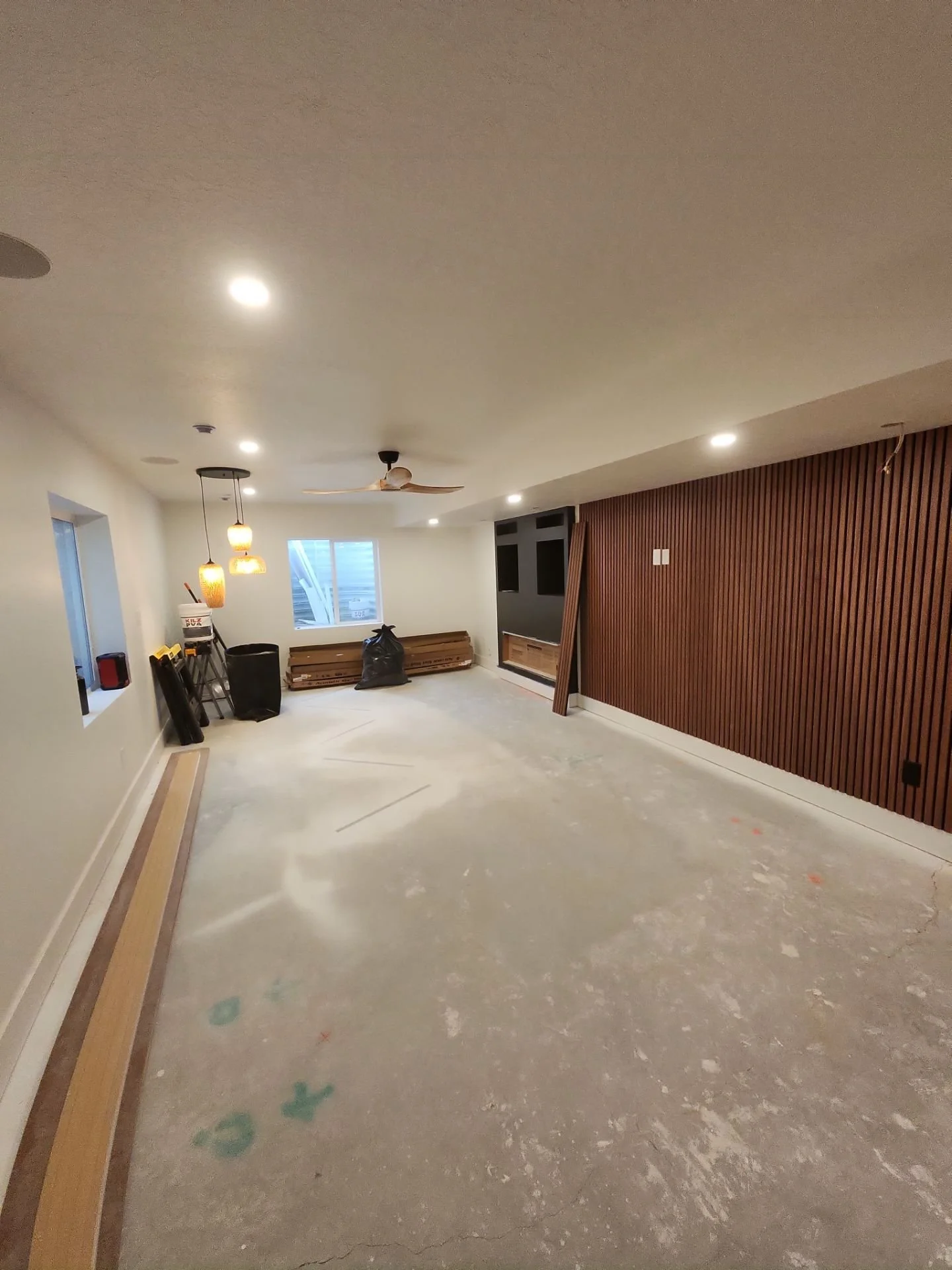 Unfinished basement room with construction materials, wood paneling, pendant lights, a ceiling fan, and a window.