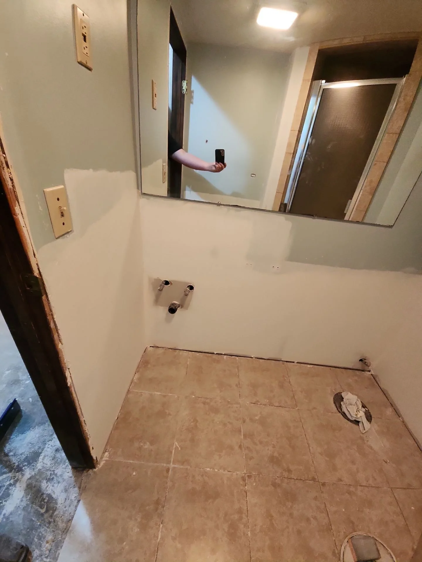 Empty bathroom with exposed plumbing for a sink and a drain on the floor, partially painted walls, a large mirror reflecting on the wall, and construction tools on the floor.
