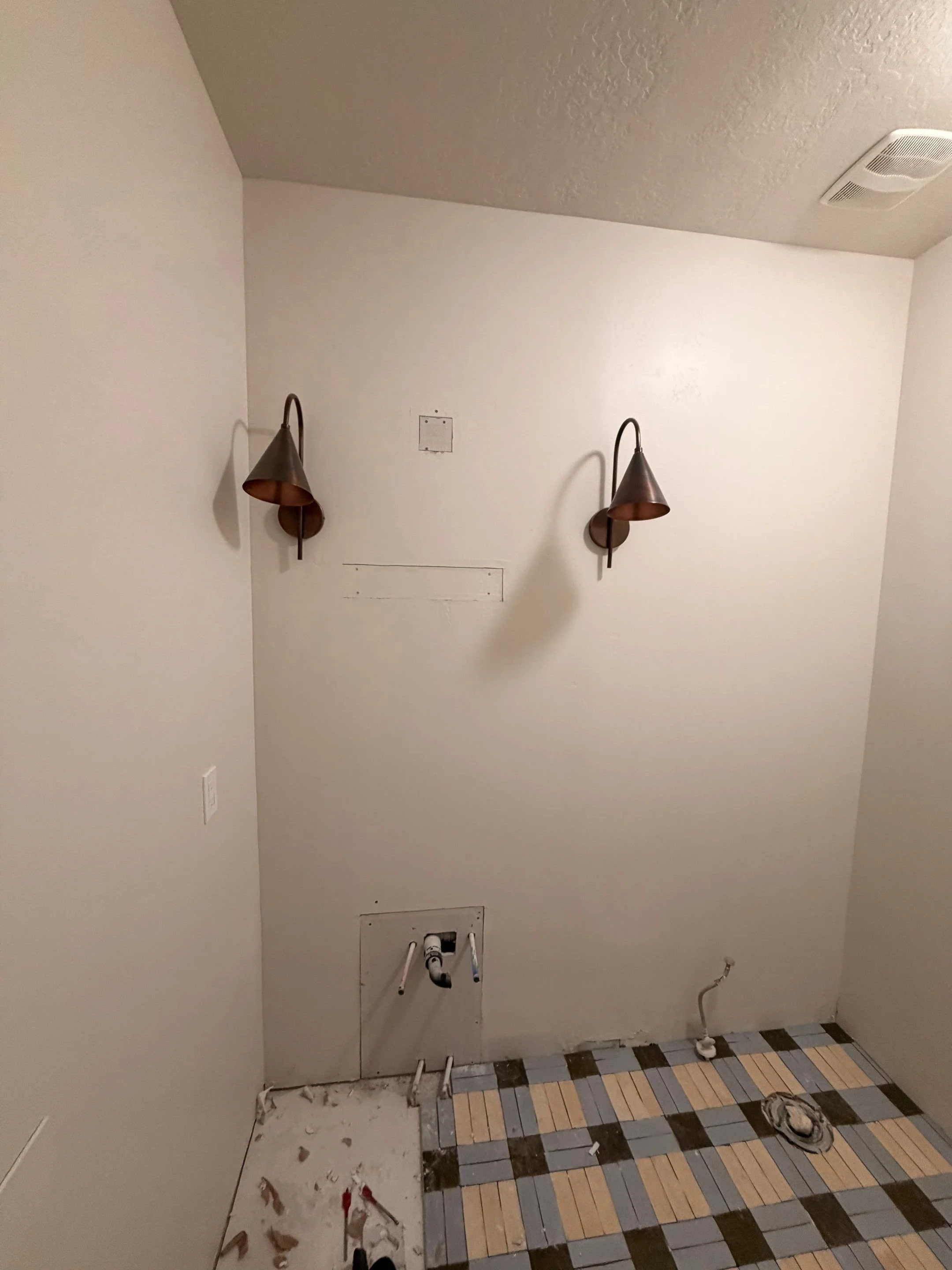 Empty bathroom with two wall-mounted light fixtures, a vent on the ceiling, and a partially installed floor with brown, blue, and beige tiles. Plumbing pipes are visible on the wall and floor, indicating ongoing renovation.
