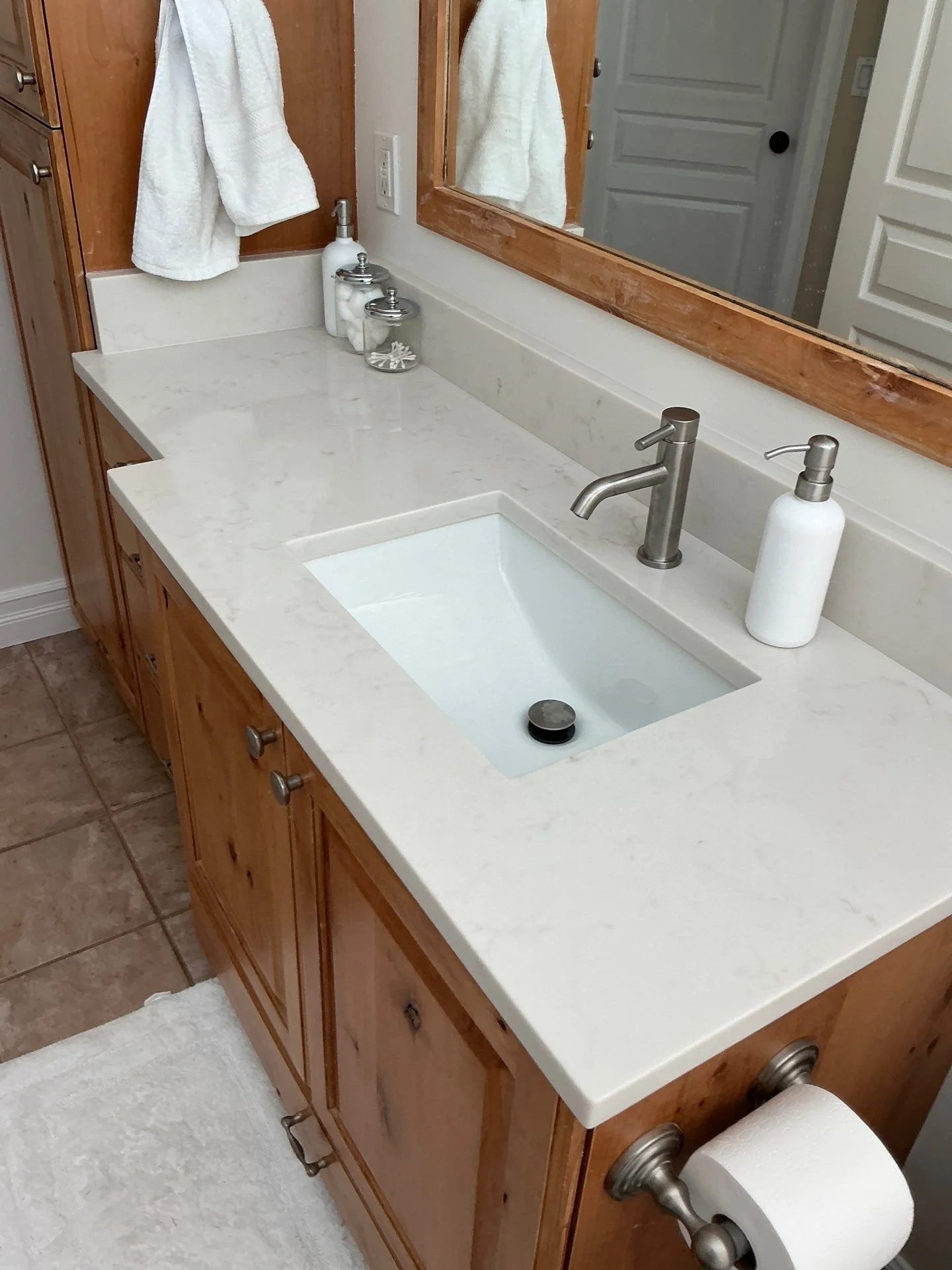 Bathroom vanity with a white marble countertop, under-mount sink, brushed metal faucet, mirror framed in wood, white towels, soap dispenser, toothbrush holder, and a toilet paper roll.