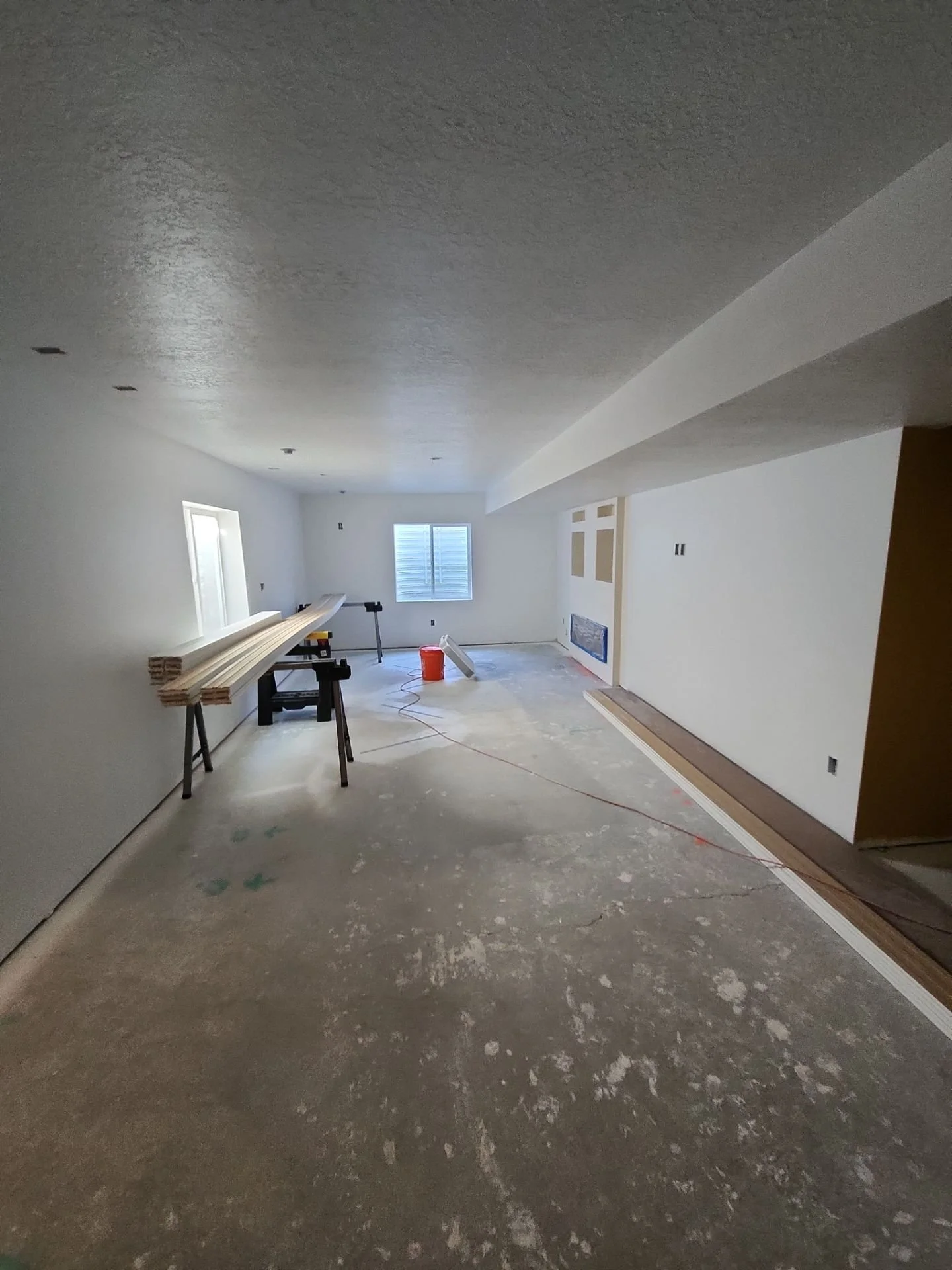 A room under construction with unfinished flooring, construction tools, and building materials like wood planks on tables, with windows and white walls.