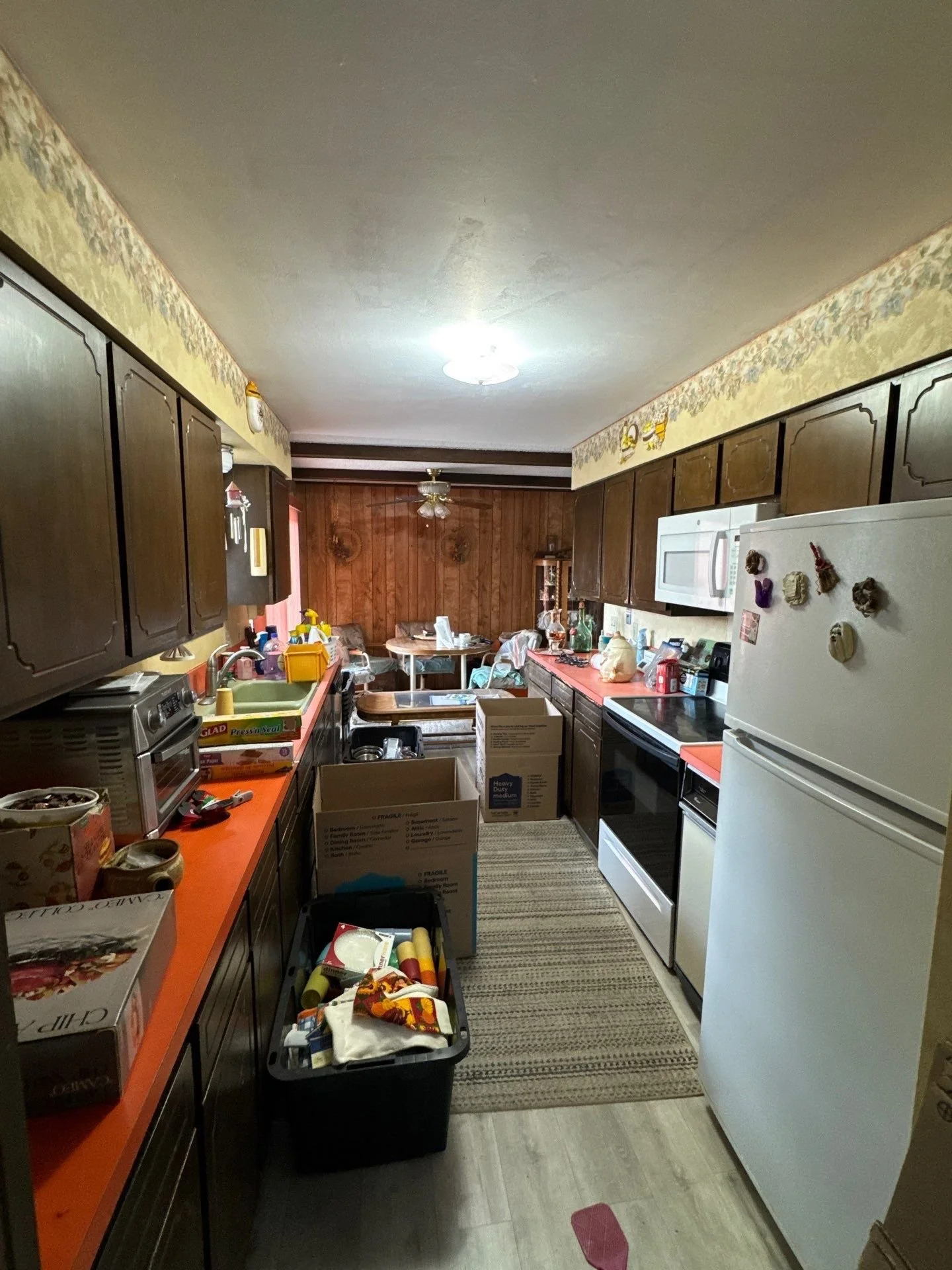 A cluttered kitchen with wooden cabinets, an orange countertop, and a white refrigerator. There are boxes and various items on the counters and the floor. A small rug is in the center, and a dining area with a round table and chairs is visible at the