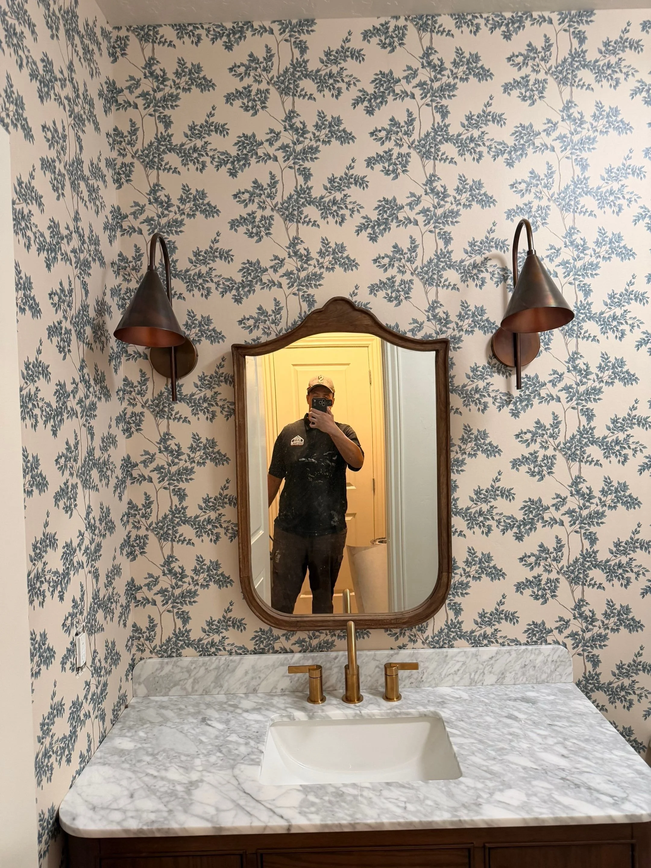 Bathroom with blue leaf-patterned wallpaper, a marble countertop with a sink, gold fixtures, and a wooden framed mirror. Two wall-mounted bronze lamps are beside the mirror. A person taking a selfie is visible in the mirror.