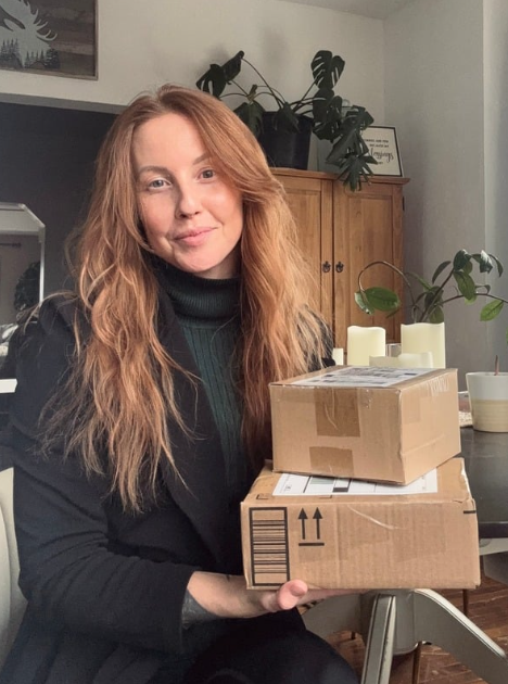 A woman with long red hair and a black turtleneck sweater smiling while holding two cardboard boxes in a room with a wooden cabinet, green plants, and candles on a table.
