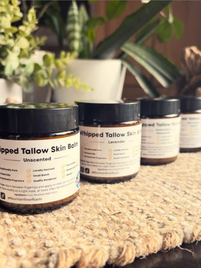 Four jars of whipped tallow skin balm labeled 'Unscented' and 'Lavender' on a woven surface, with potted plants in the background.