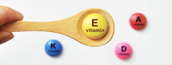 A wooden spoon holding a yellow pill labeled 'E vitamin' with three other vitamin pills nearby: blue K, pink D, and red A.