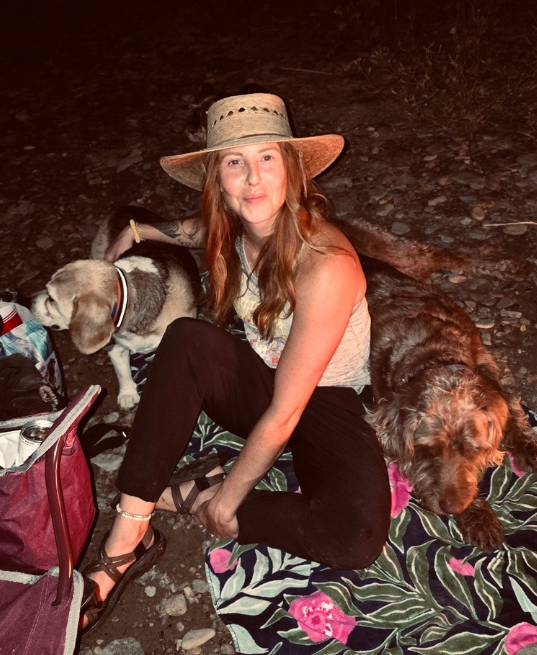 Woman wearing a straw hat sitting on a blanket with two dogs at night.