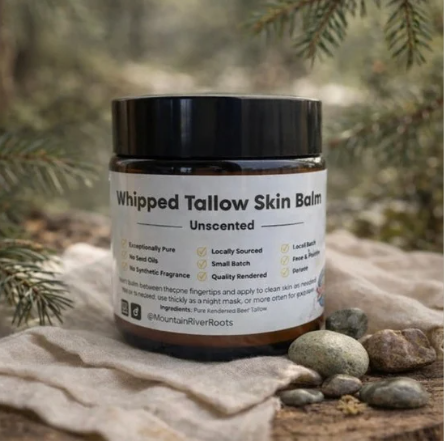 A jar of whipped tallow skin balm labeled 'Unscented' on a rocky outdoor surface with pine needles in the background.