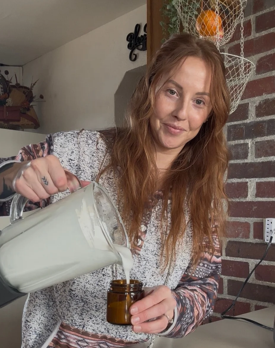 A woman with long wavy red hair pouring tallow  from a glass pitcher into a brown jar