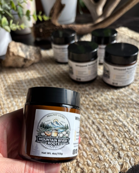 A hand holding a small brown jar labeled 'Mountain River Roots' supplement with black lid, with several similar jars on a woven surface in the background.