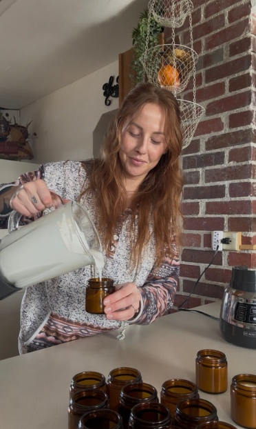 A woman pouring a liquid from a glass pitcher into small amber jars on a kitchen counter.