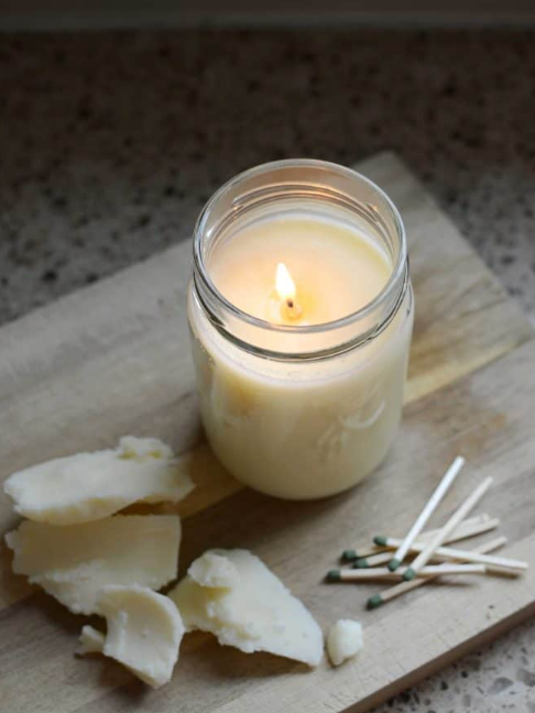 A lit white tallow candle in a glass jar, with pieces of broken tallow and several matchsticks on a wooden surface.