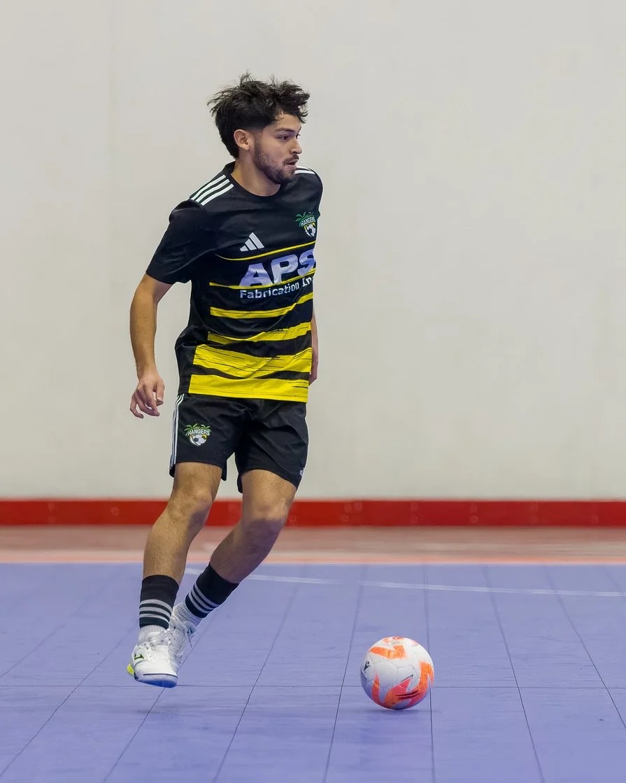 A young male soccer player with dark, tousled hair wearing a black and yellow striped jersey, black shorts, and striped socks, is controlling a white and orange soccer ball on an indoor court with a red border and a plain off-white wall in the backgr