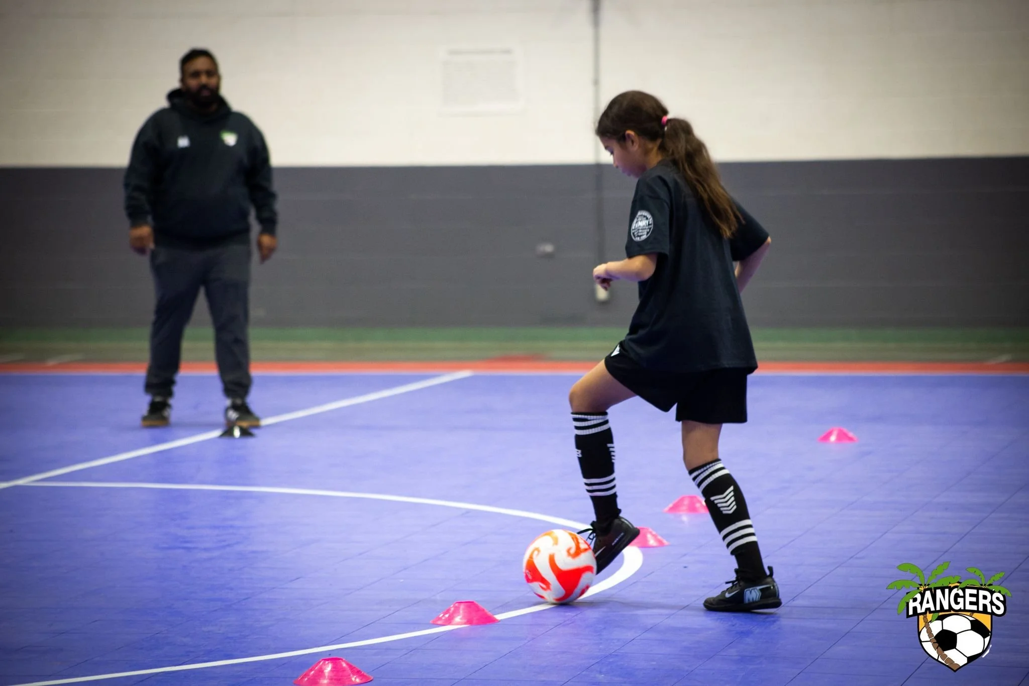 Young girl in black sports uniform practicing soccer drills on an indoor court with orange cones, supervised by an adult coach in the background, with a soccer team logo on the bottom right corner.
