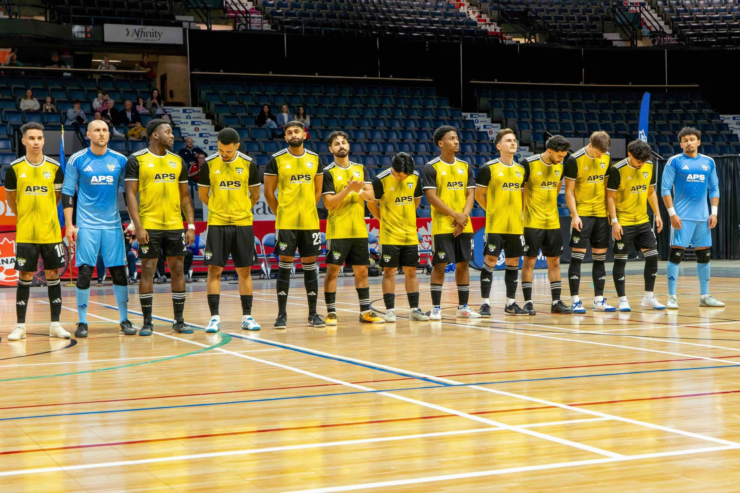 A group of indoor soccer players standing in a line on a court, wearing yellow and black uniforms with two players in light blue goalkeeper uniforms, in an arena with empty seats and some spectators in the background.