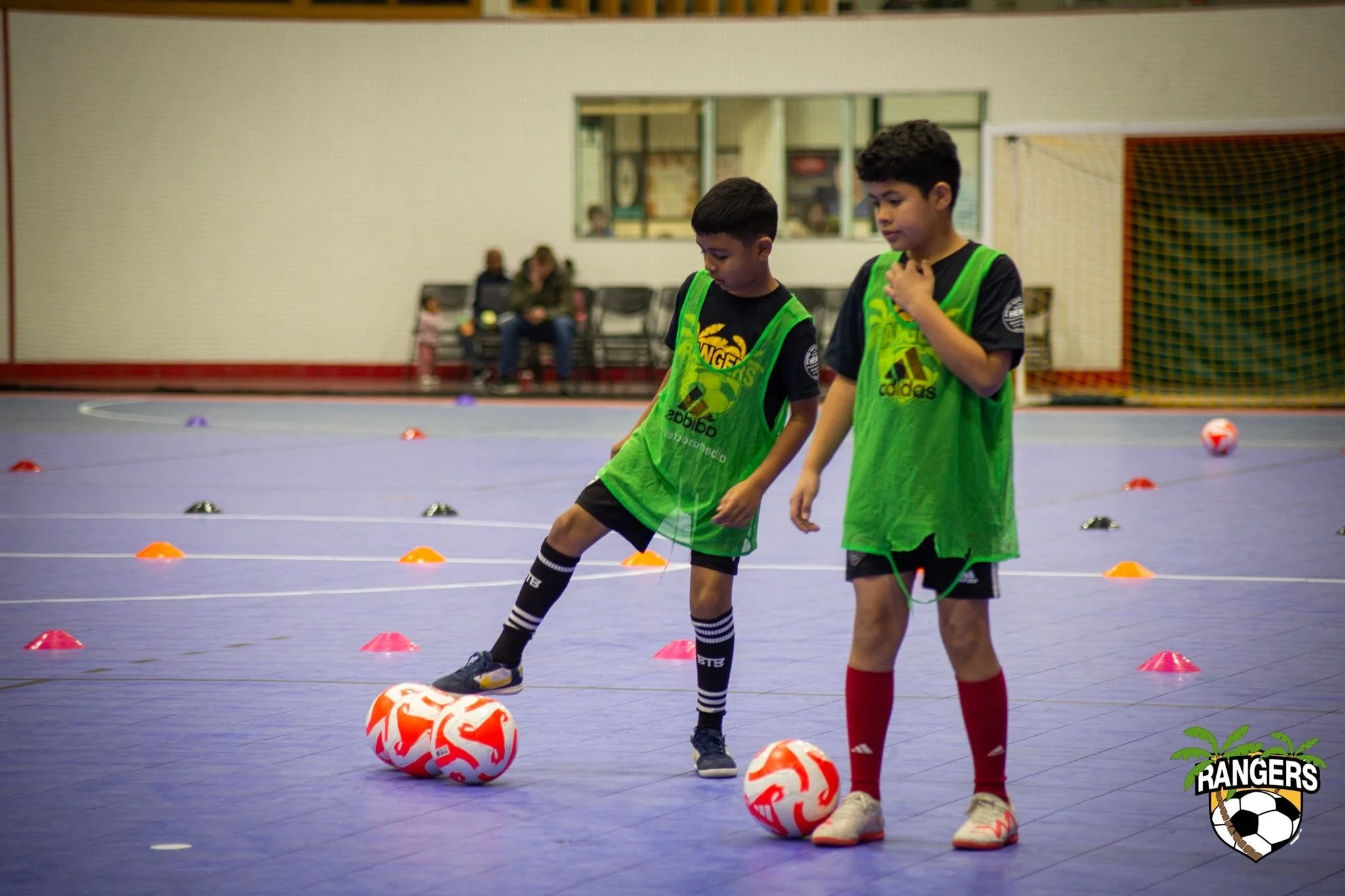 Two young boys in green sports bibs practicing indoor soccer dribbling with orange and white soccer balls on a blue court, with orange cones set up for drills, and a soccer goal with a net in the background.