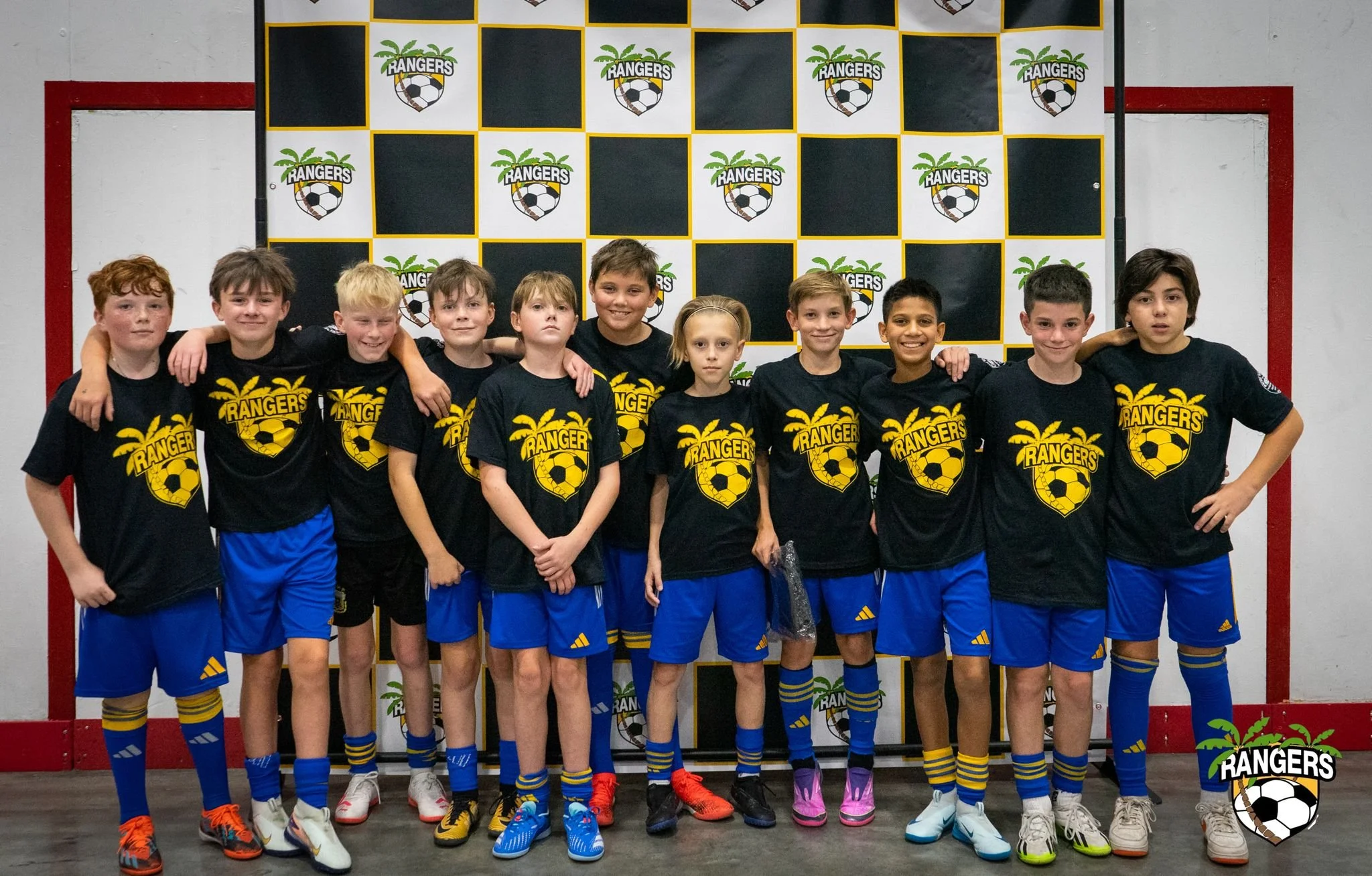 Youth soccer team in black and yellow jerseys standing together with arms around each other, posing for a group photo in front of a black and yellow checkered background with ranger logo.